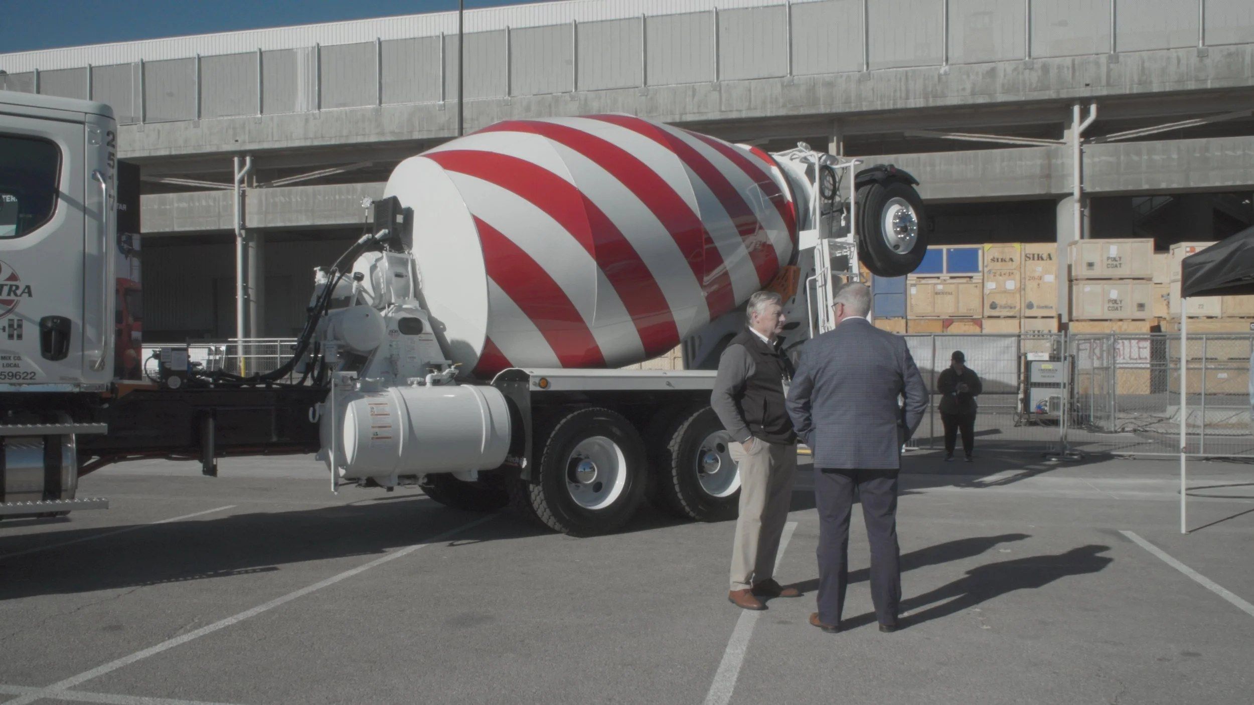 John Wilson stands near a concrete truck and another man in an official film still.