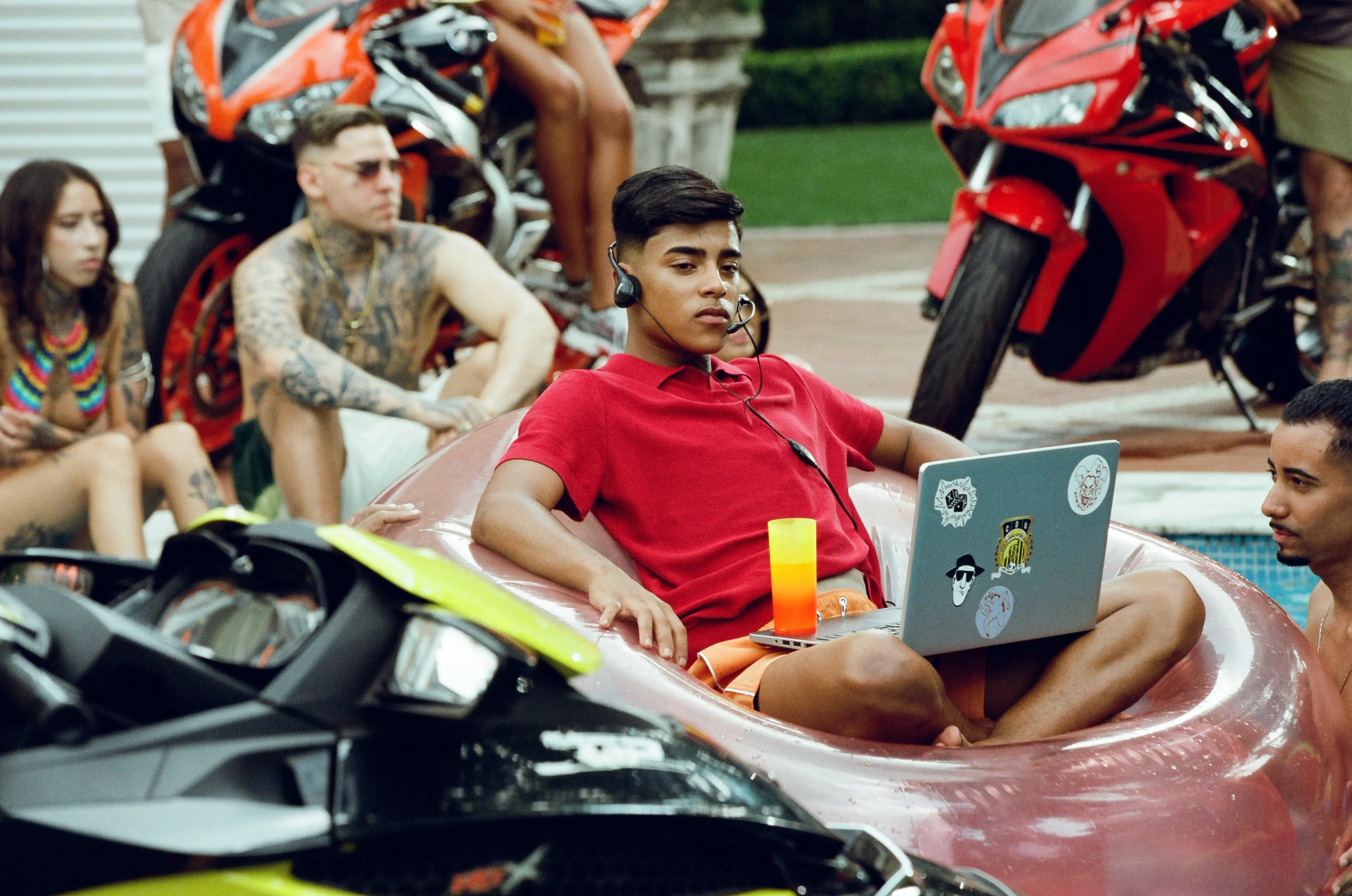 A young man in a red shirt wears a headset and works on a laptop at a pool party in a Rauls promotional image.