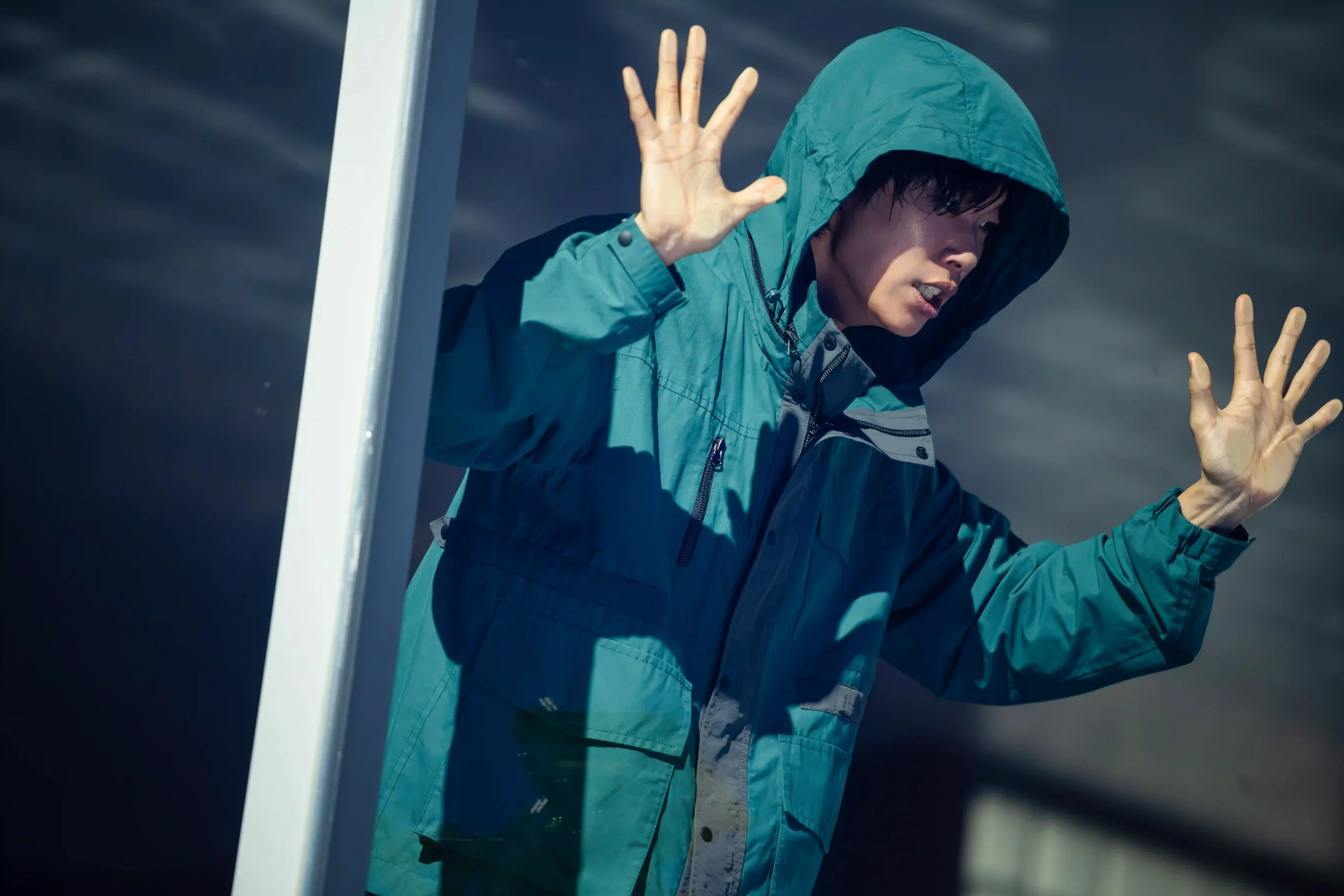 Ryu Jun-yeol’s character wears a teal hooded coat and raises both hands beside a glass door.
