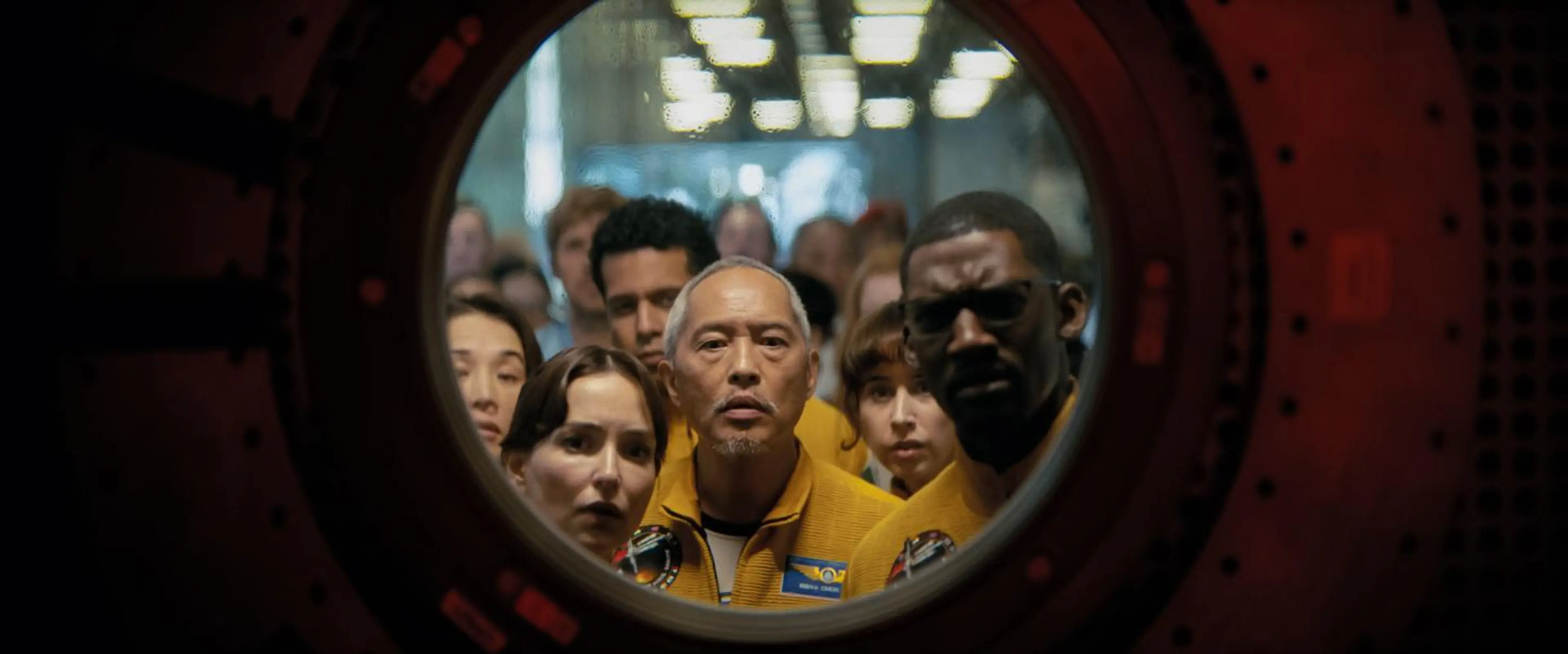 A group in yellow uniforms peers through a circular window into a spacecraft.