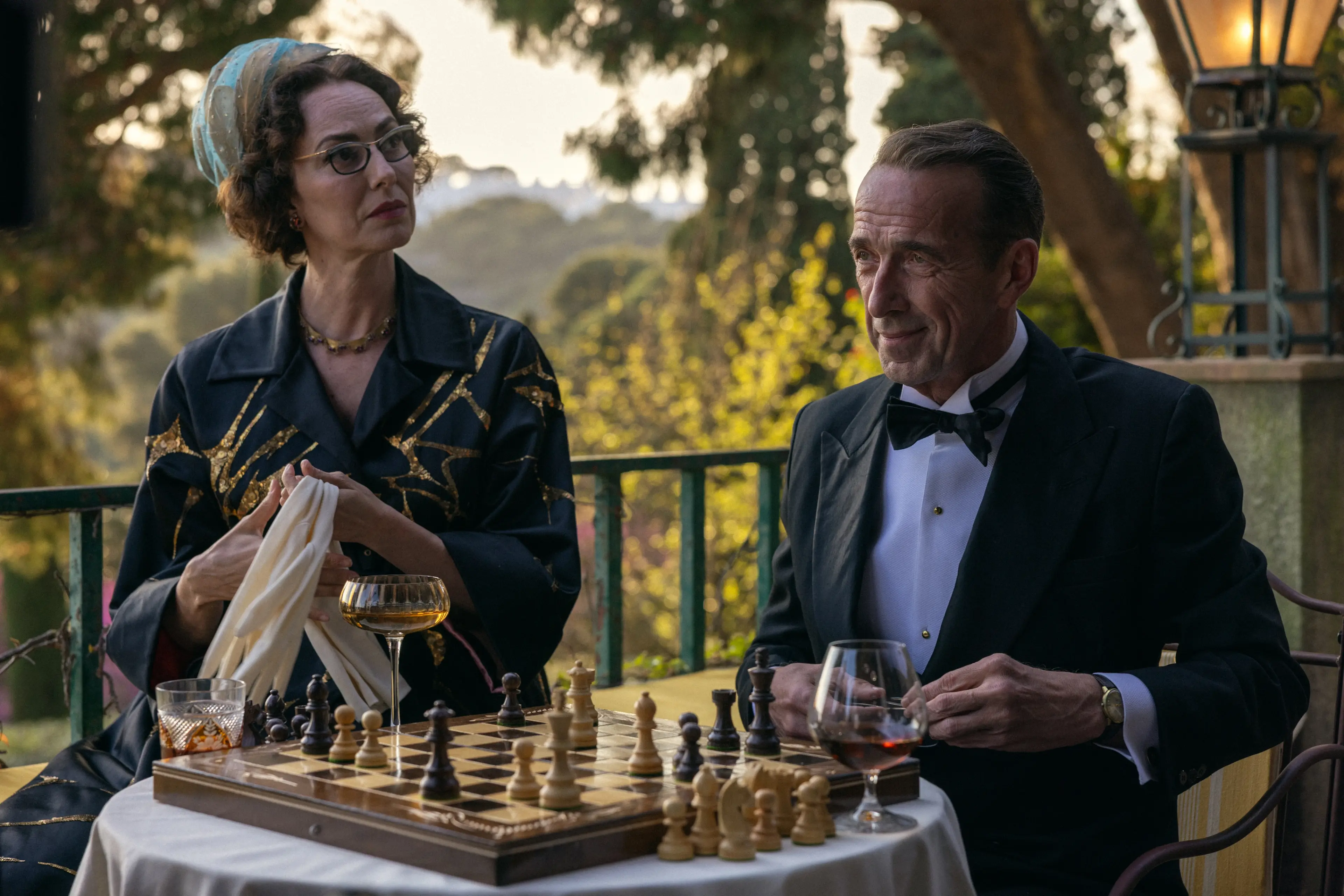 José Coronado as Basil and Maribel Verdú as Mrs. Ausländer sit beside a chessboard on the hotel terrace.