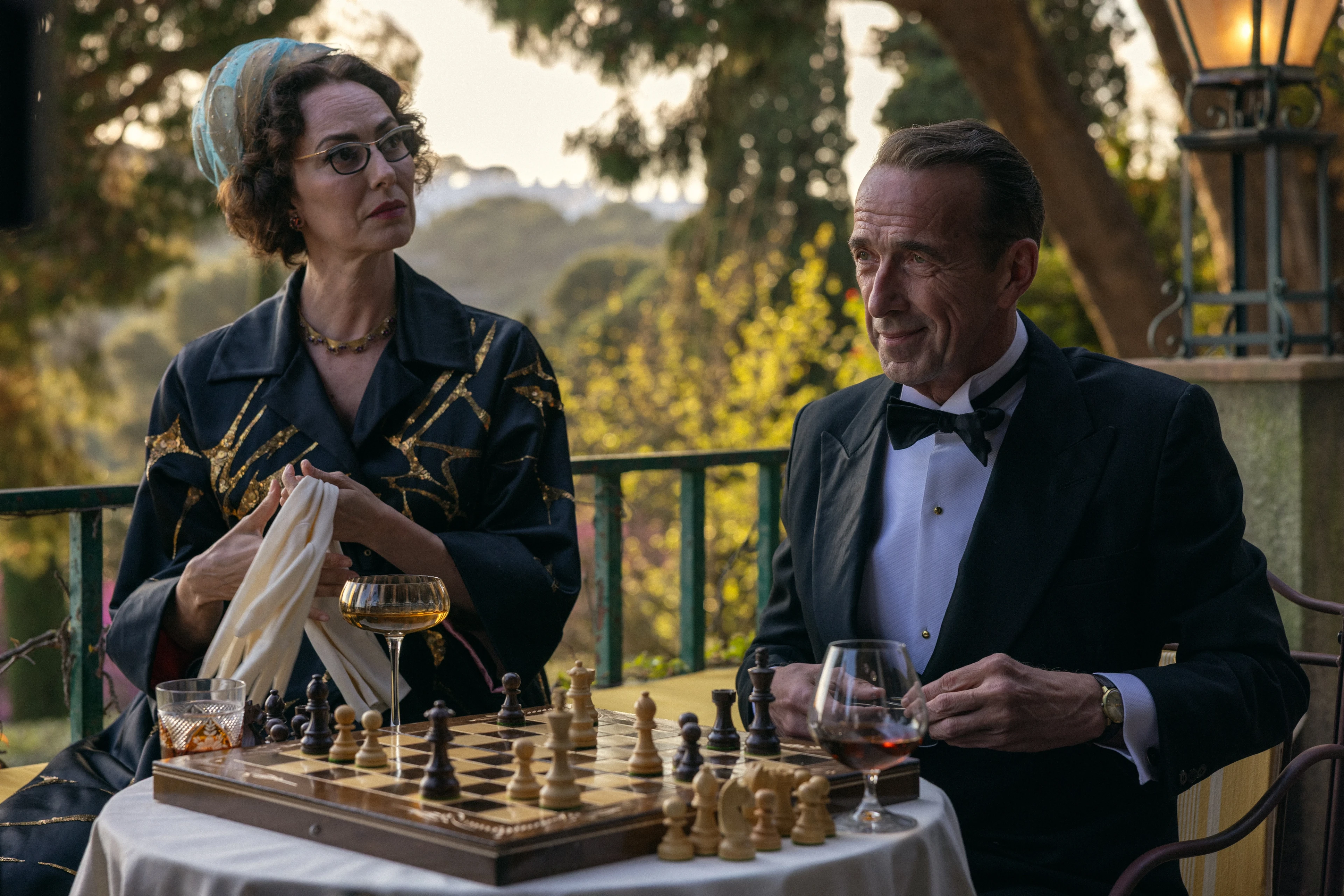 José Coronado as Basil and Maribel Verdú as Mrs. Ausländer sit beside a chessboard on the hotel terrace.
