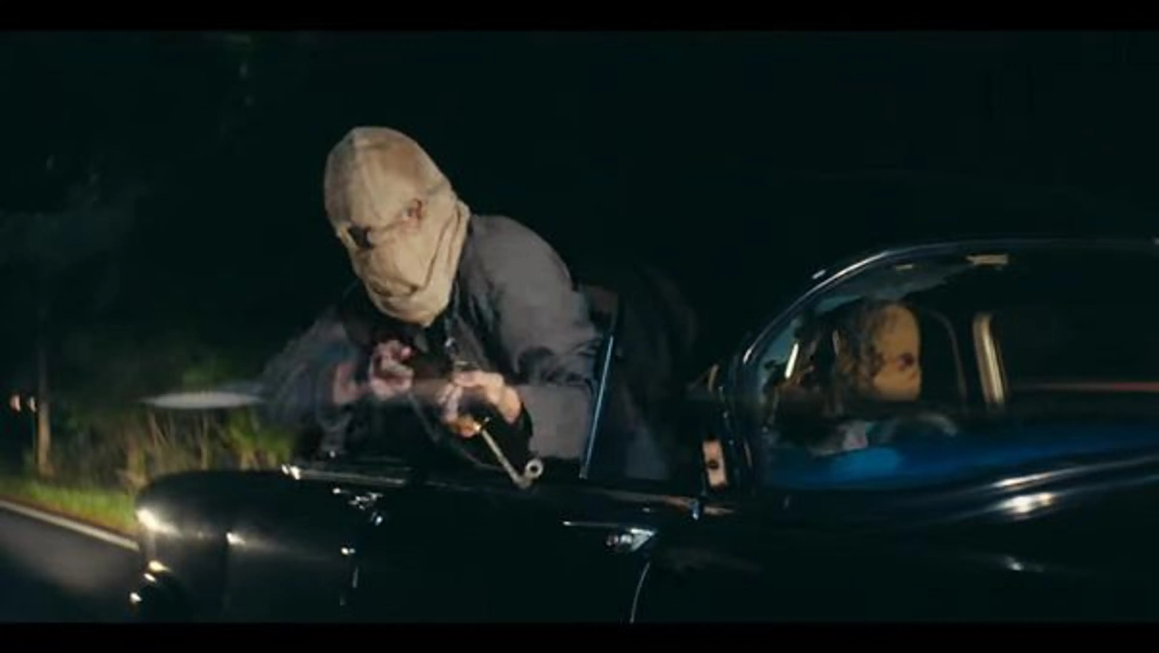 A masked gunman leans from a moving car with a firearm in By Any Means.