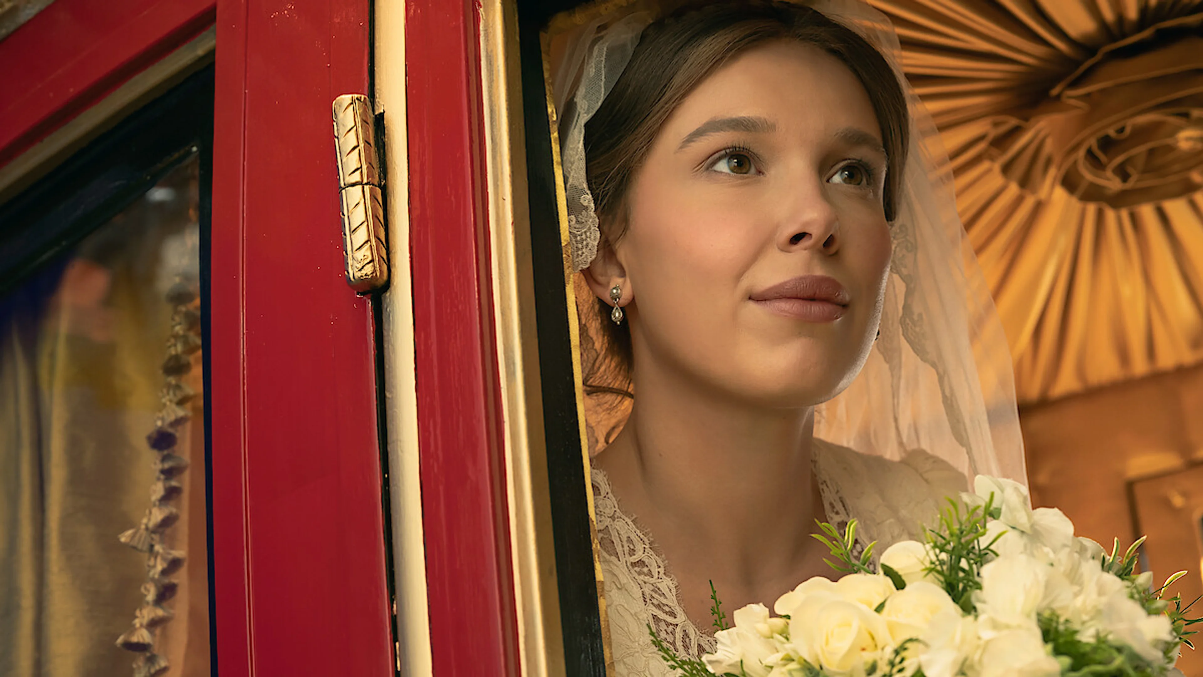 Enola wears a wedding veil and holds white flowers at the window of a red carriage.