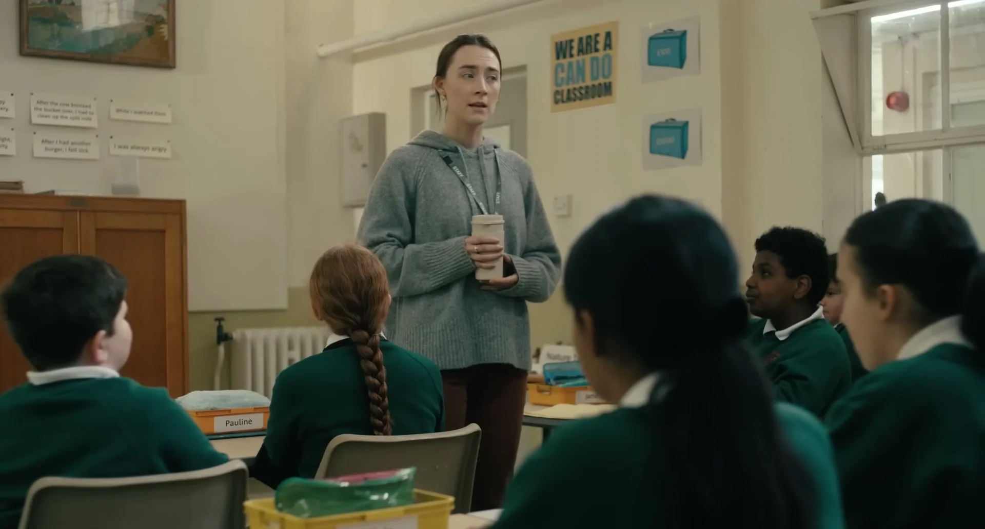 Maria holds a cup while speaking to pupils seated in her classroom.