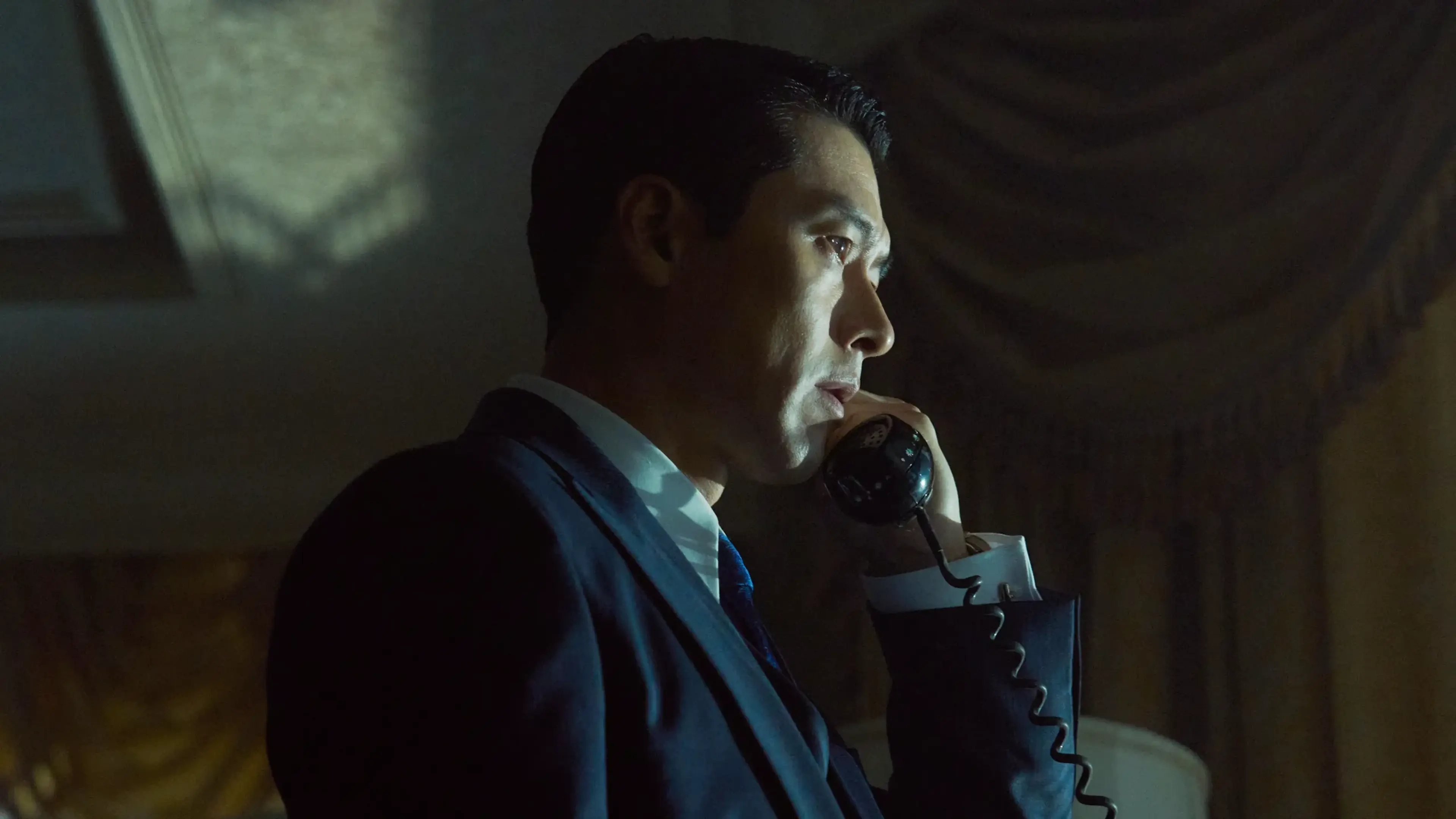 Ki-tae holds a telephone receiver to his ear while wearing a suit in a dim room.