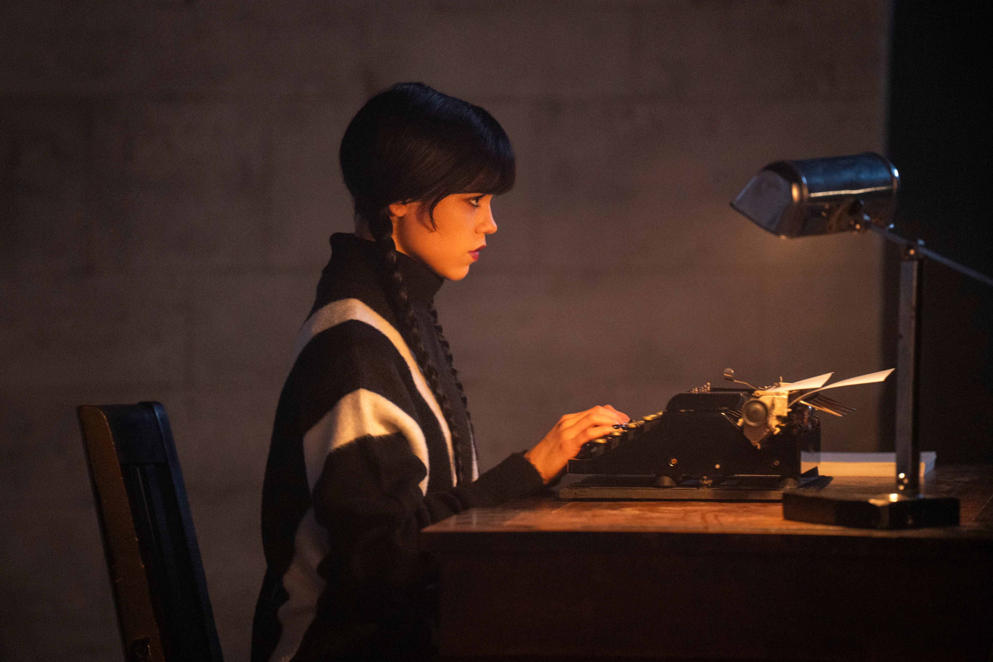 Jenna Ortega as Wednesday Addams sits at a typewriter in a Season 3 production image.