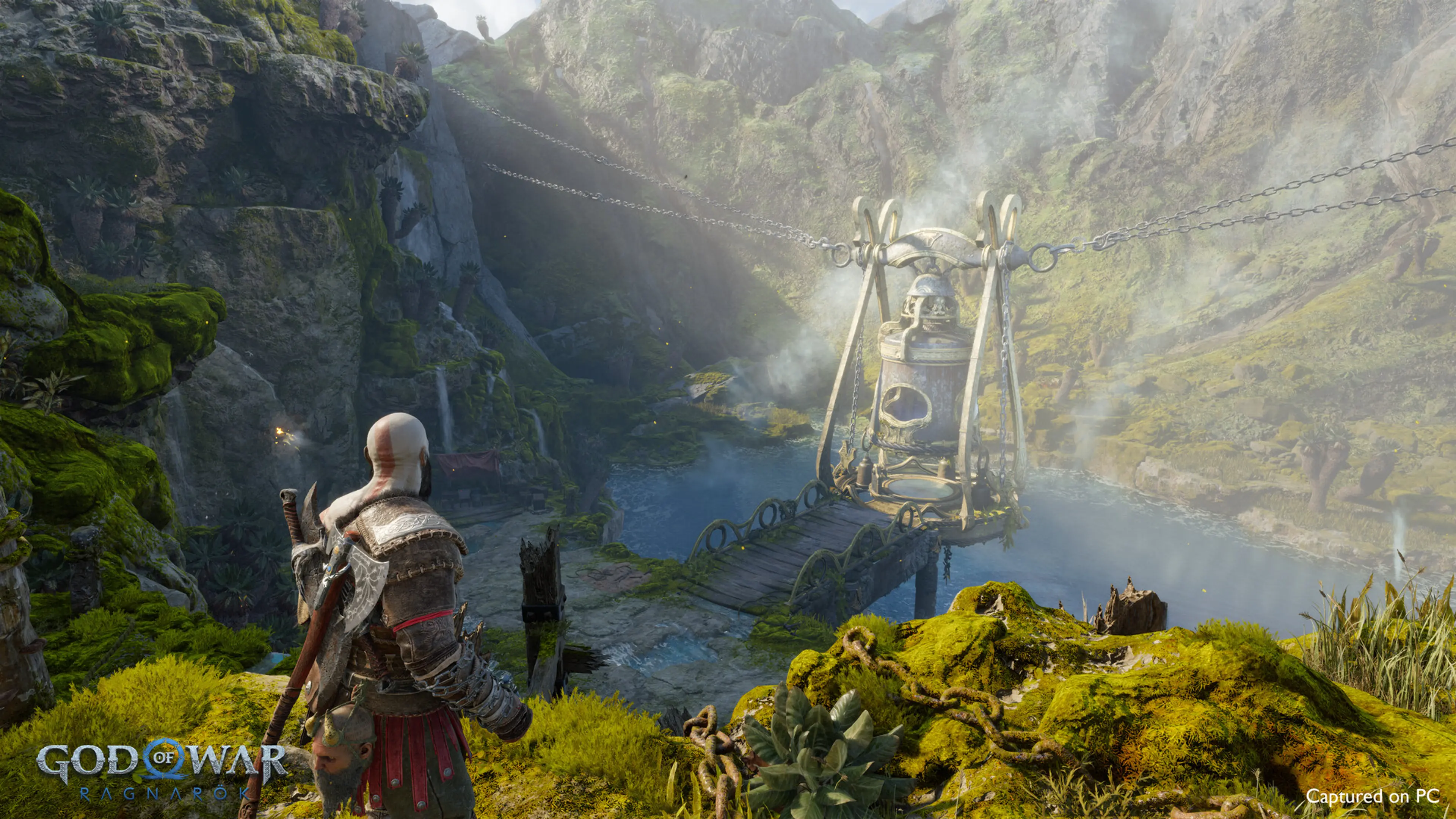 Kratos looks across a moss-covered ravine toward a large mechanism suspended over water in God of War Ragnarok.