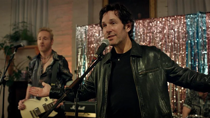 Rick spreads his arms at a microphone while a guitarist plays behind him.