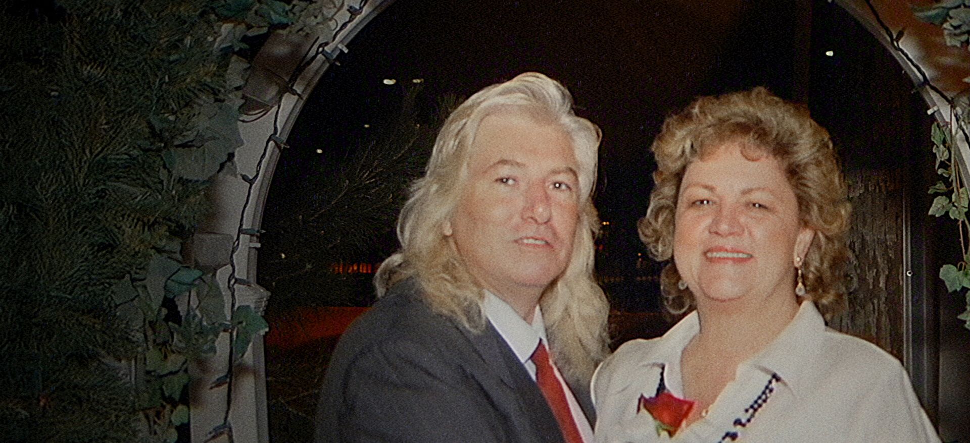 A silver-haired man in a dark suit stands beside a woman in a white blouse beneath an arch.