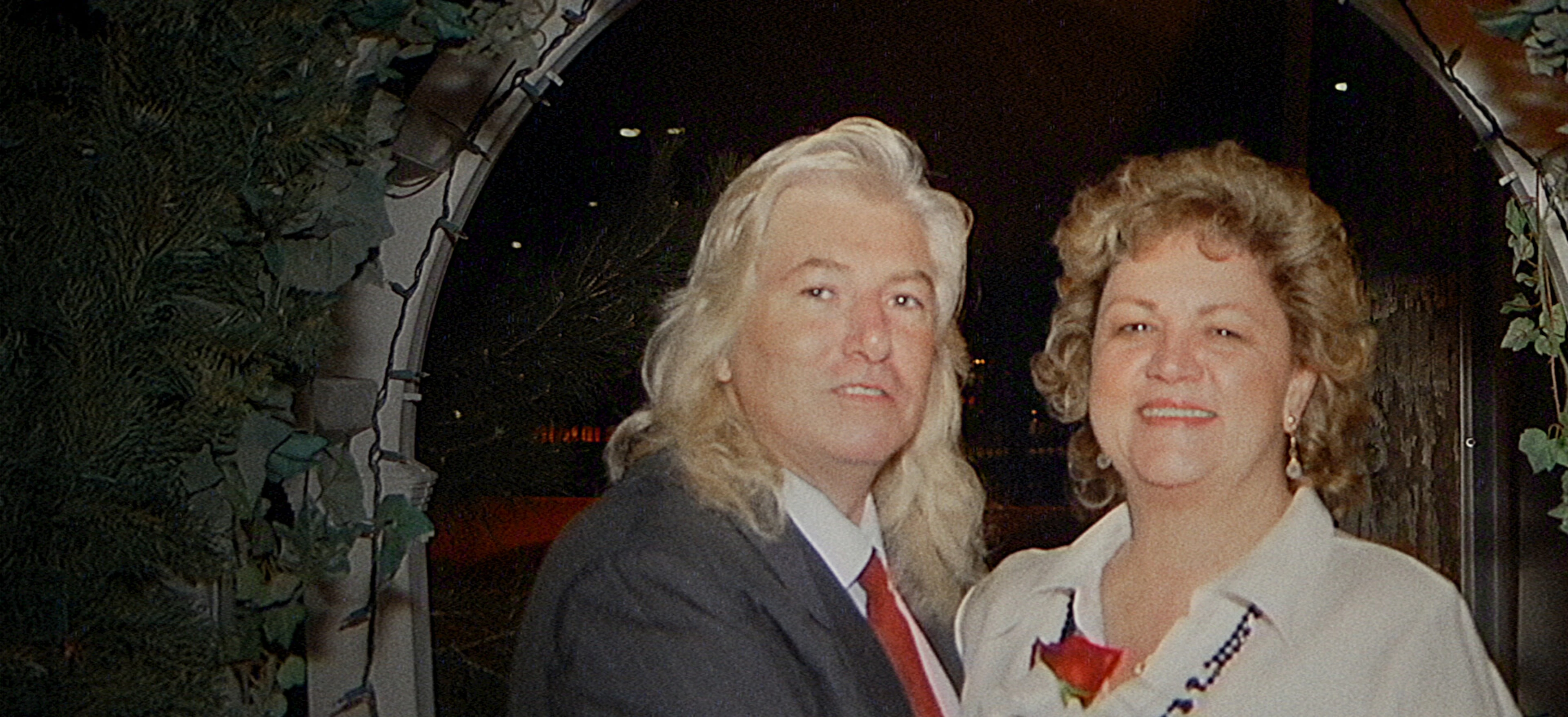 A silver-haired man in a dark suit stands beside a woman in a white blouse beneath an arch.
