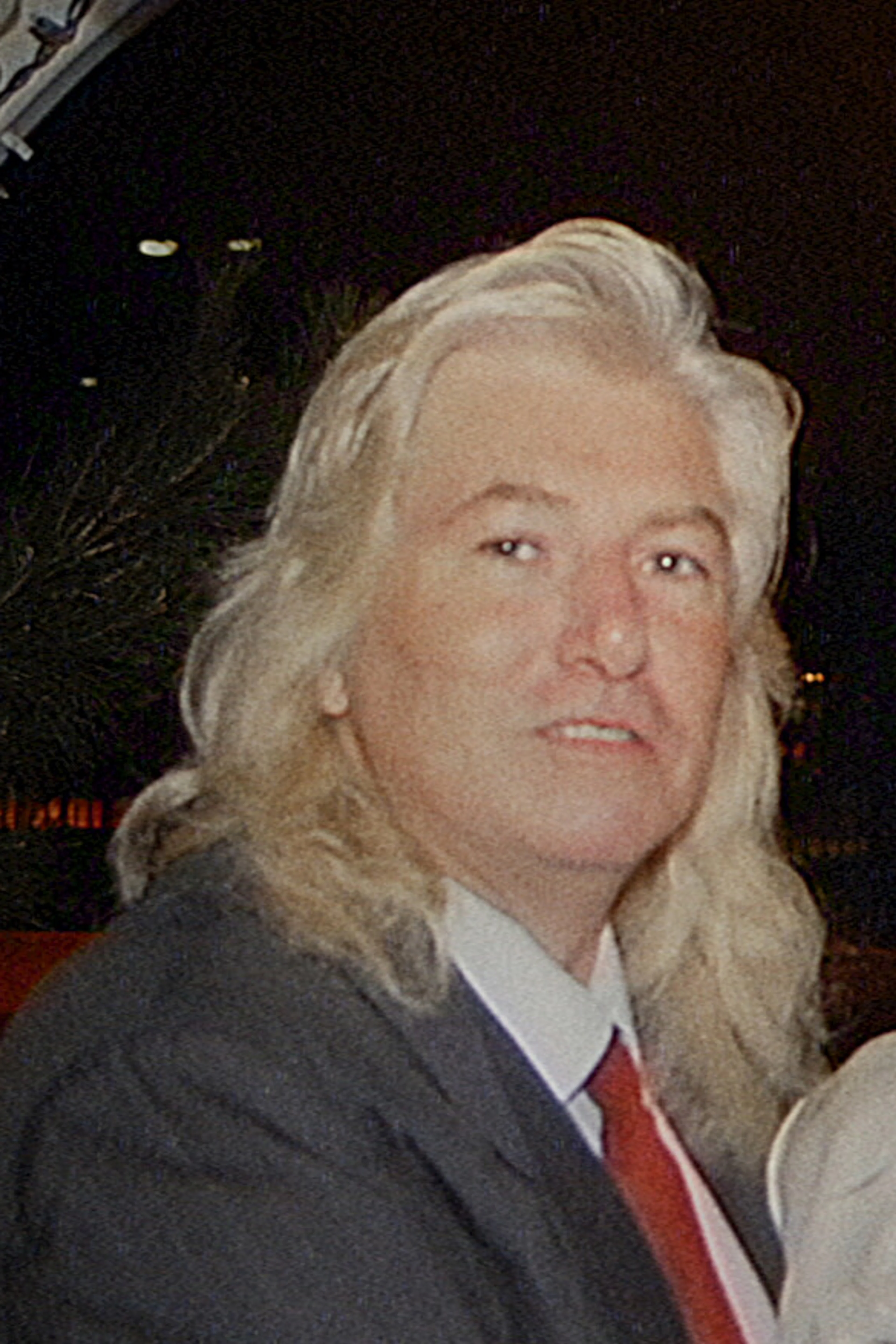 A silver-haired man in a dark suit stands beside a woman in a white blouse beneath an arch.