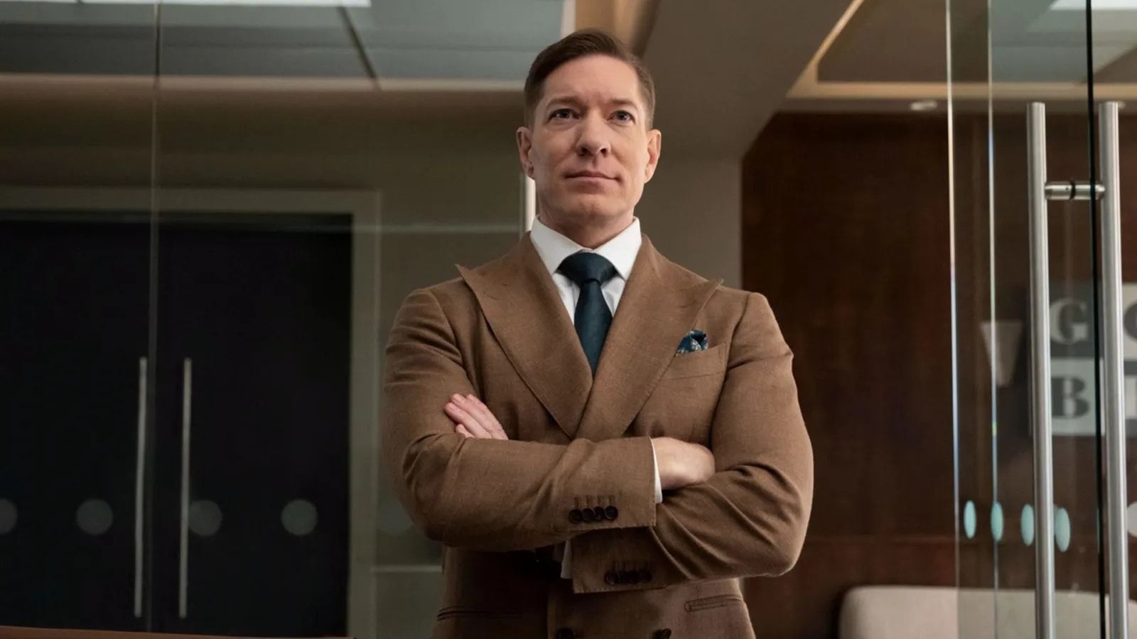 Joseph Sikora as Bill Sterling stands in an office in a brown suit.