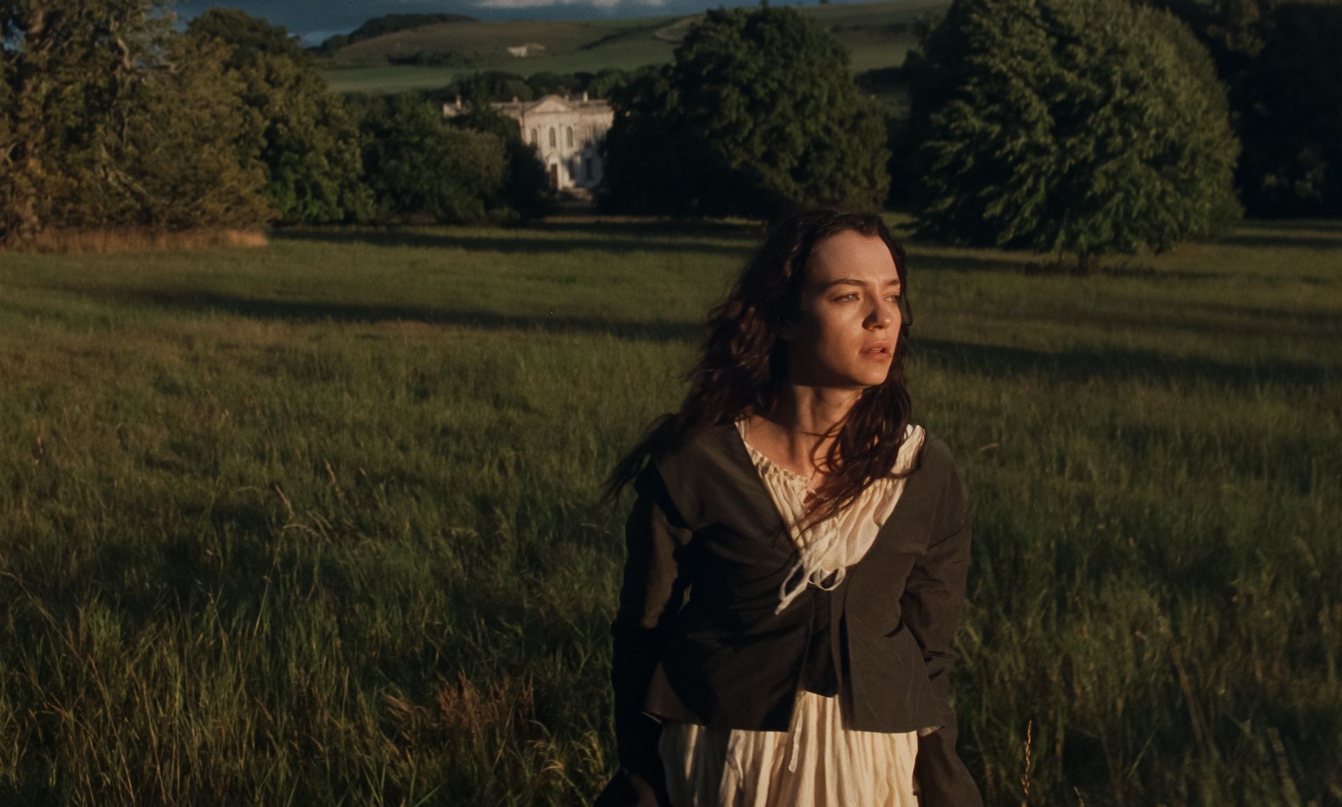 Marianne stands in long grass with a large country house behind her.