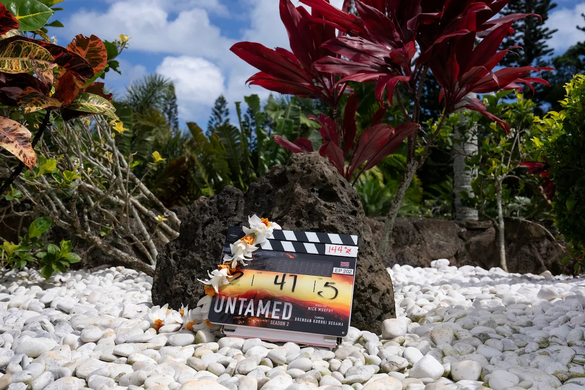 A clapperboard labeled UNTAMED rests on volcanic rock among tropical plants.