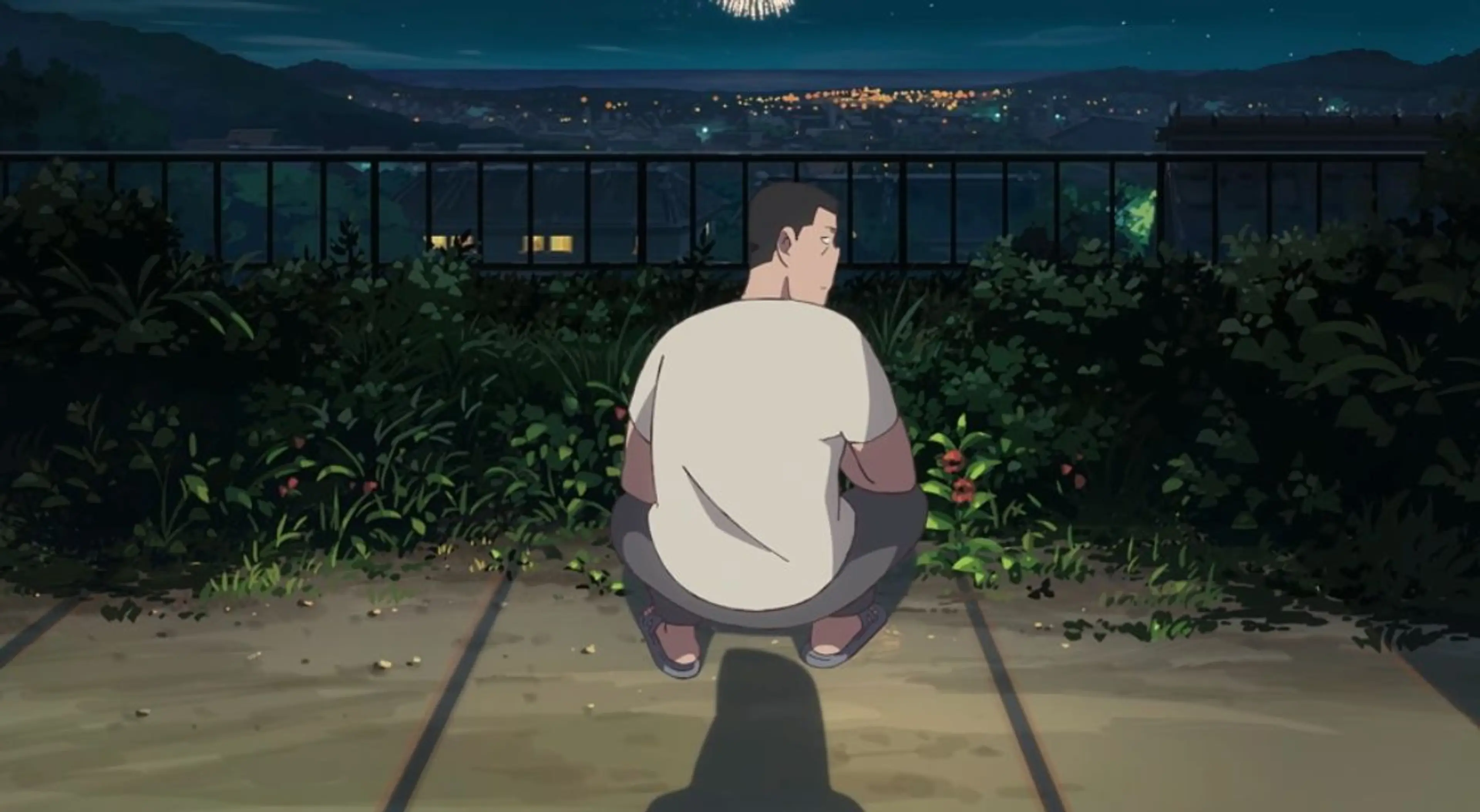 Akutsu crouches beside plants on a balcony as fireworks light up the town beyond it.