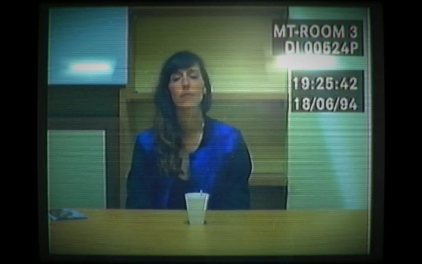 A woman wearing a blue jacket sits at a police interview table; the top-right corner of the footage includes a recording date and time.