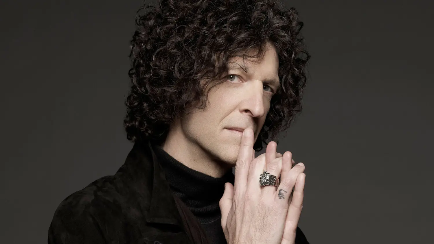Howard Stern poses against a dark studio backdrop with his hands raised near his face.