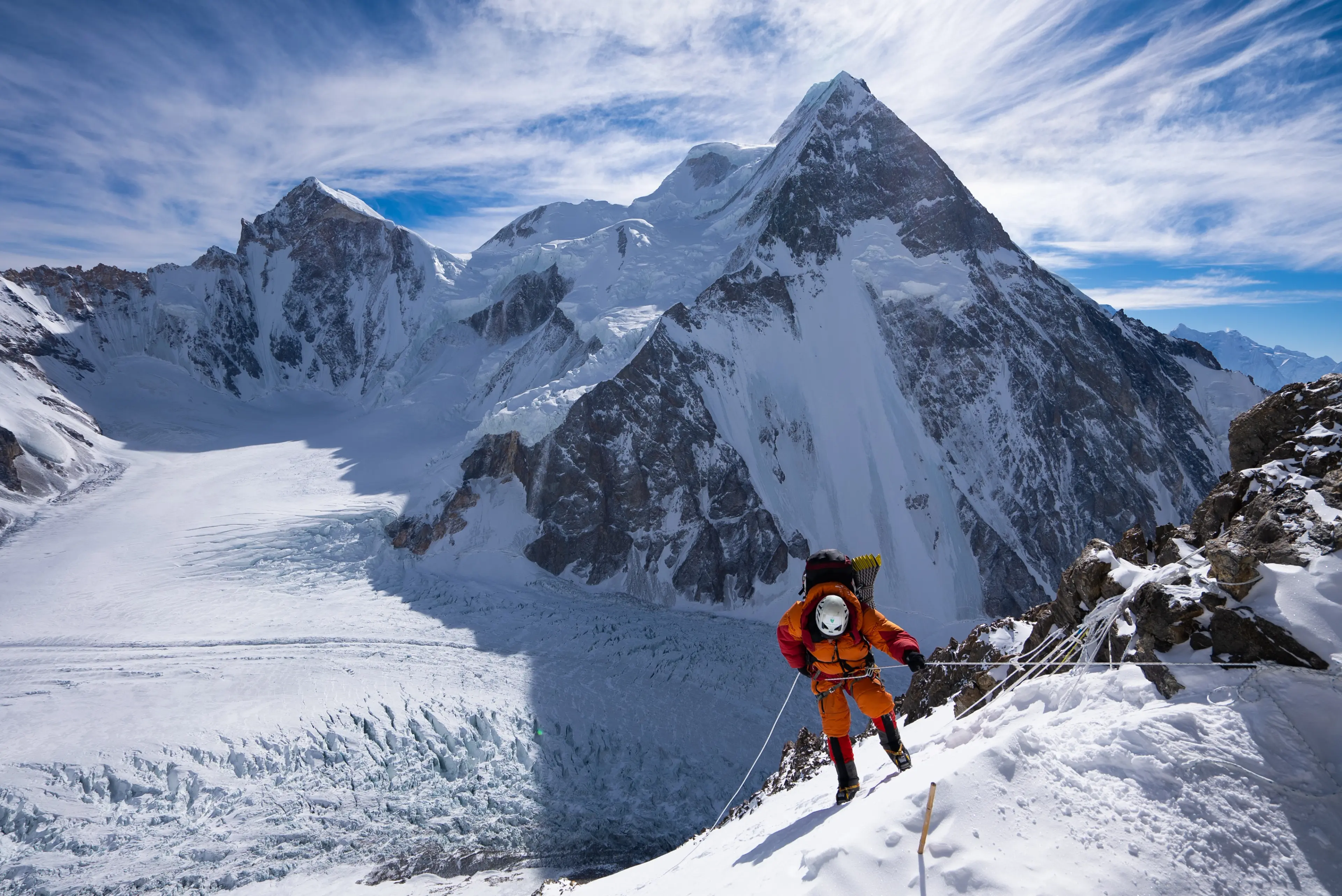 A climber in an orange suit moves up a fixed rope above a vast snowy glacier.