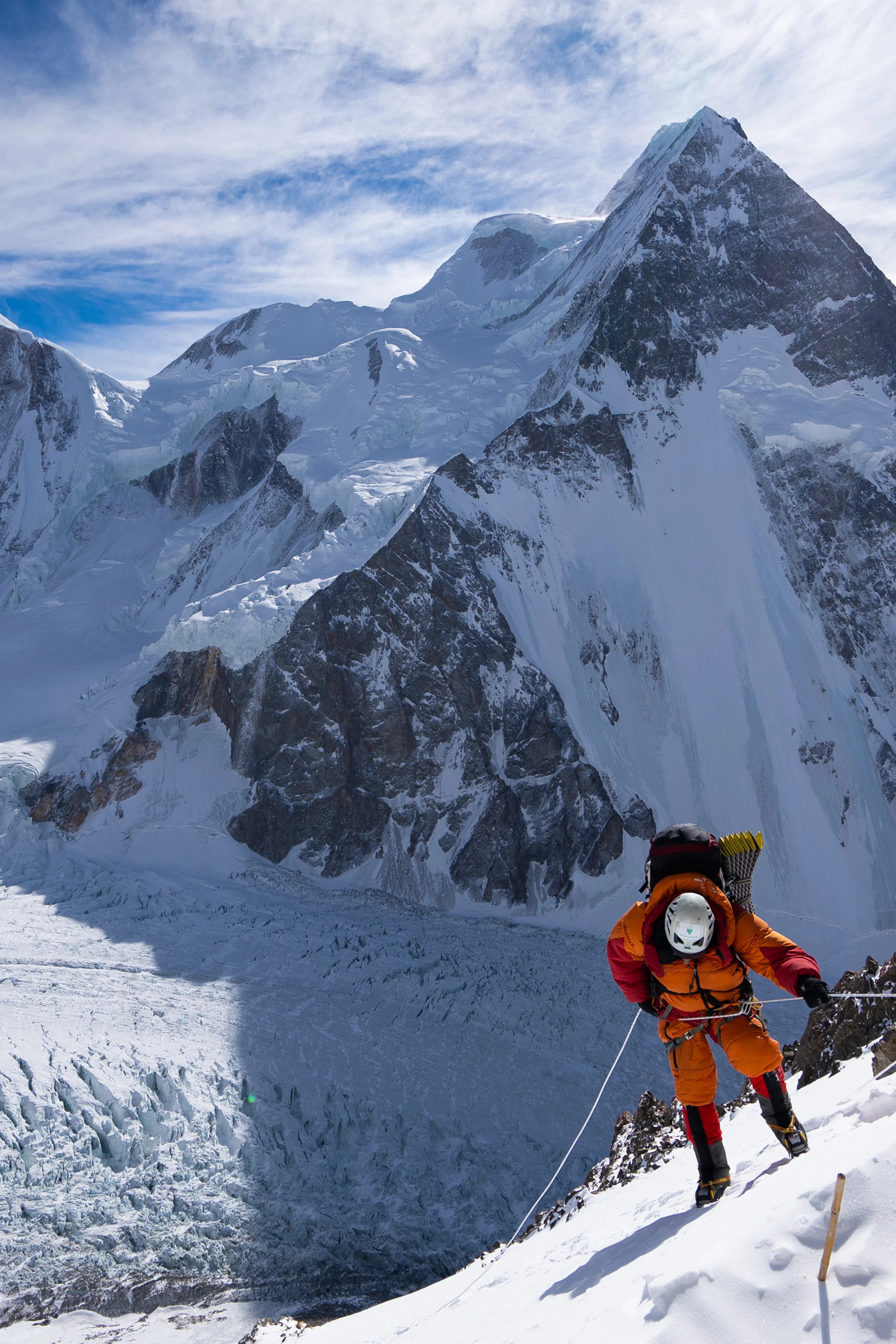 A climber in an orange suit moves up a fixed rope above a vast snowy glacier.