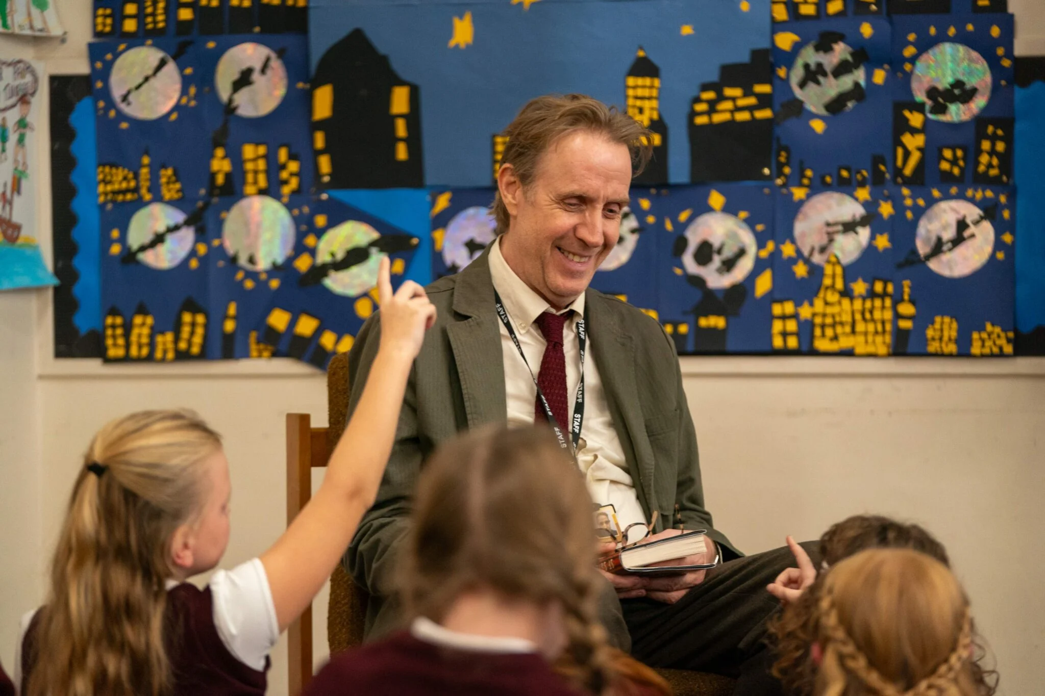 Rhys Ifans as Tony smiles while talking with pupils in a primary-school classroom.