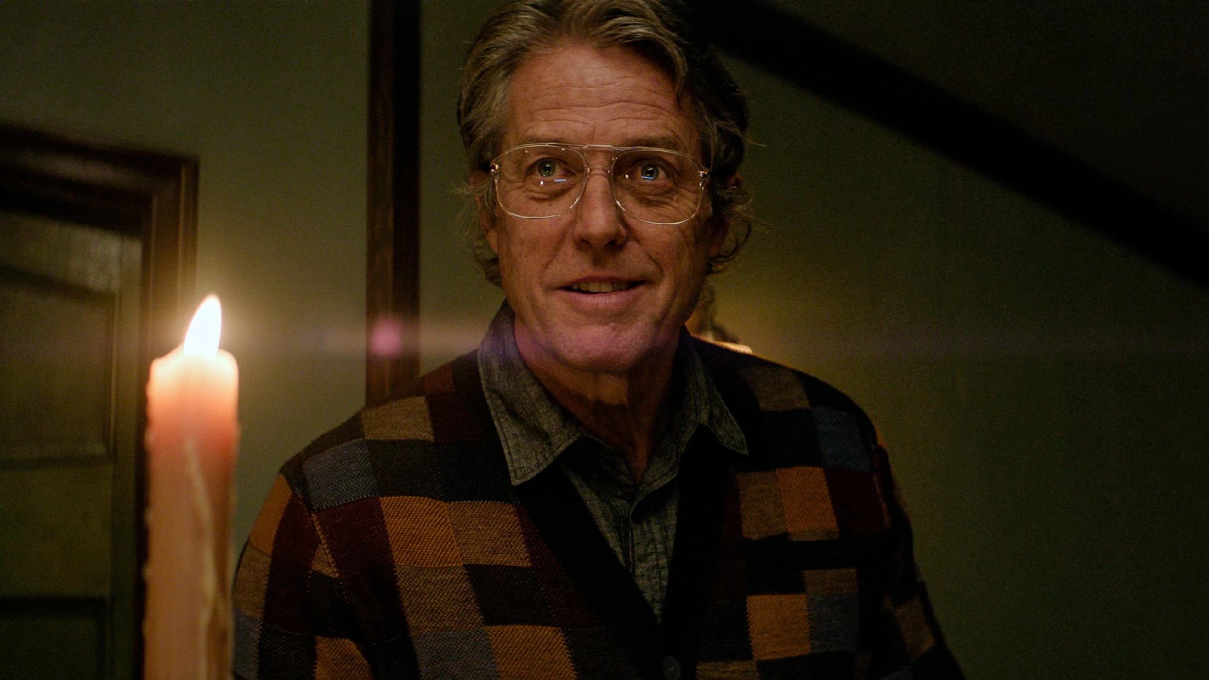 Hugh Grant as Mr. Reed holds a lit candle while wearing glasses and a checked cardigan.