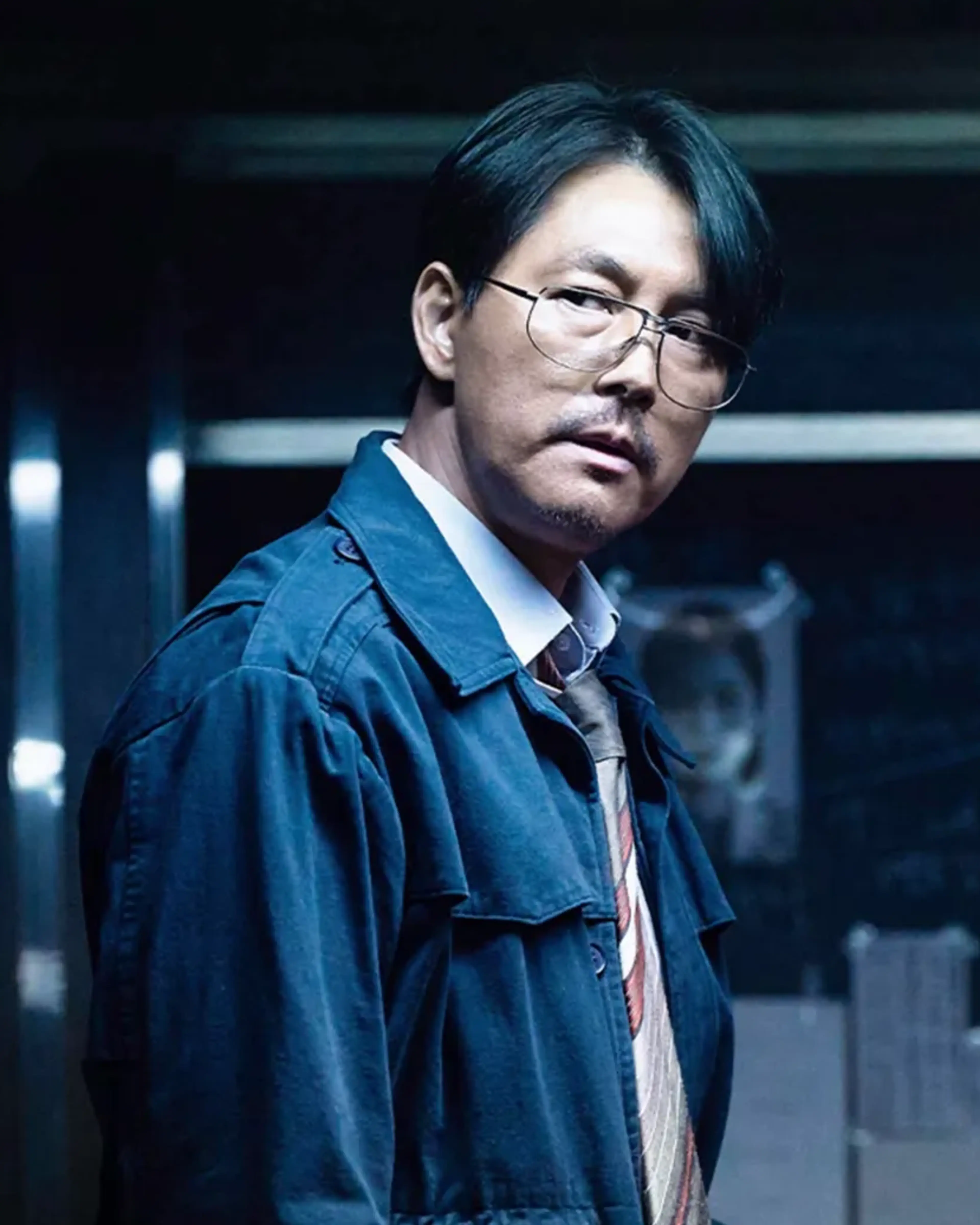 Jung Woo-sung as Geon-young stands in a dark room beside a board of documents and photographs.