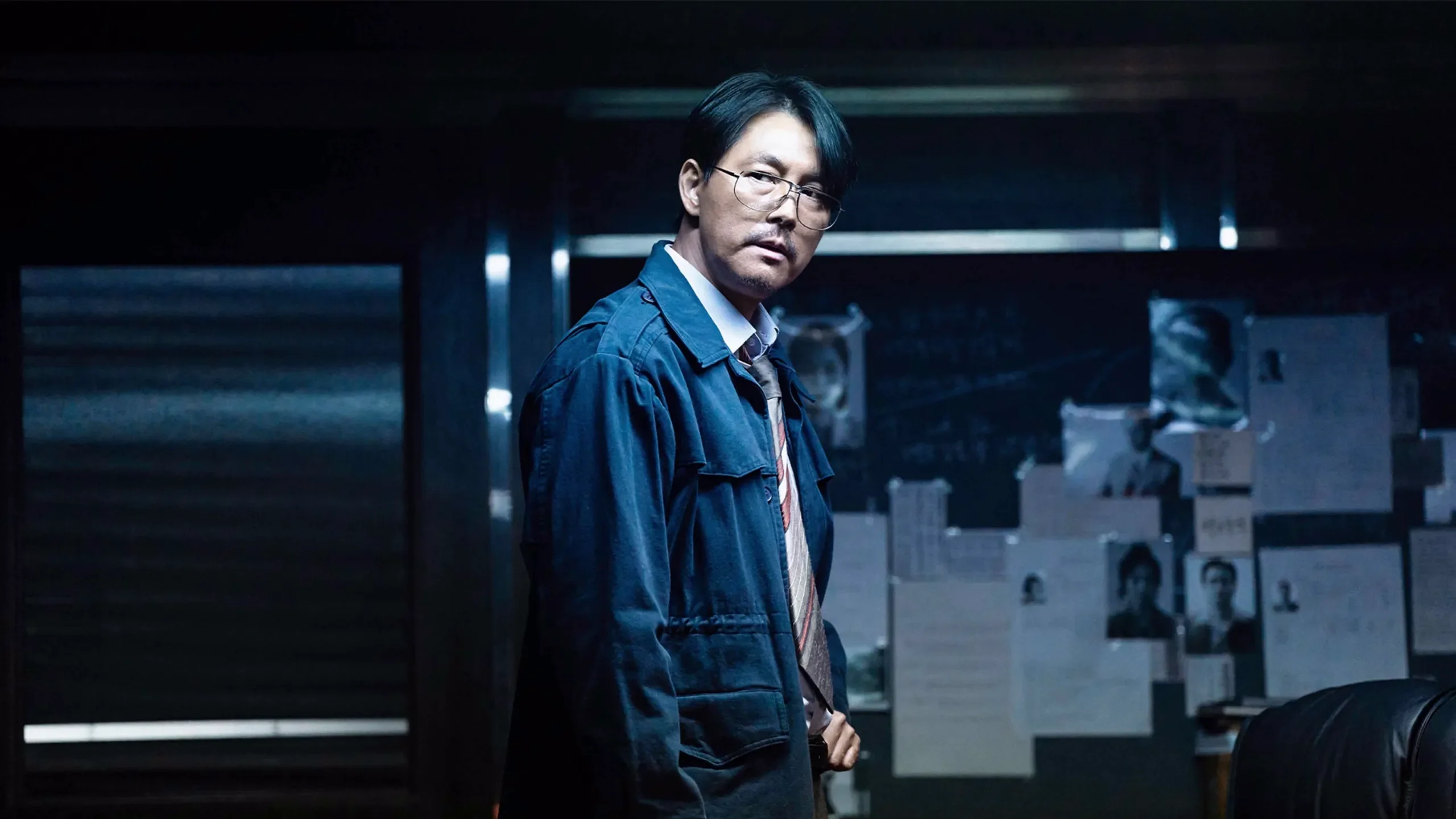 Jung Woo-sung as Geon-young stands in a dark room beside a board of documents and photographs.