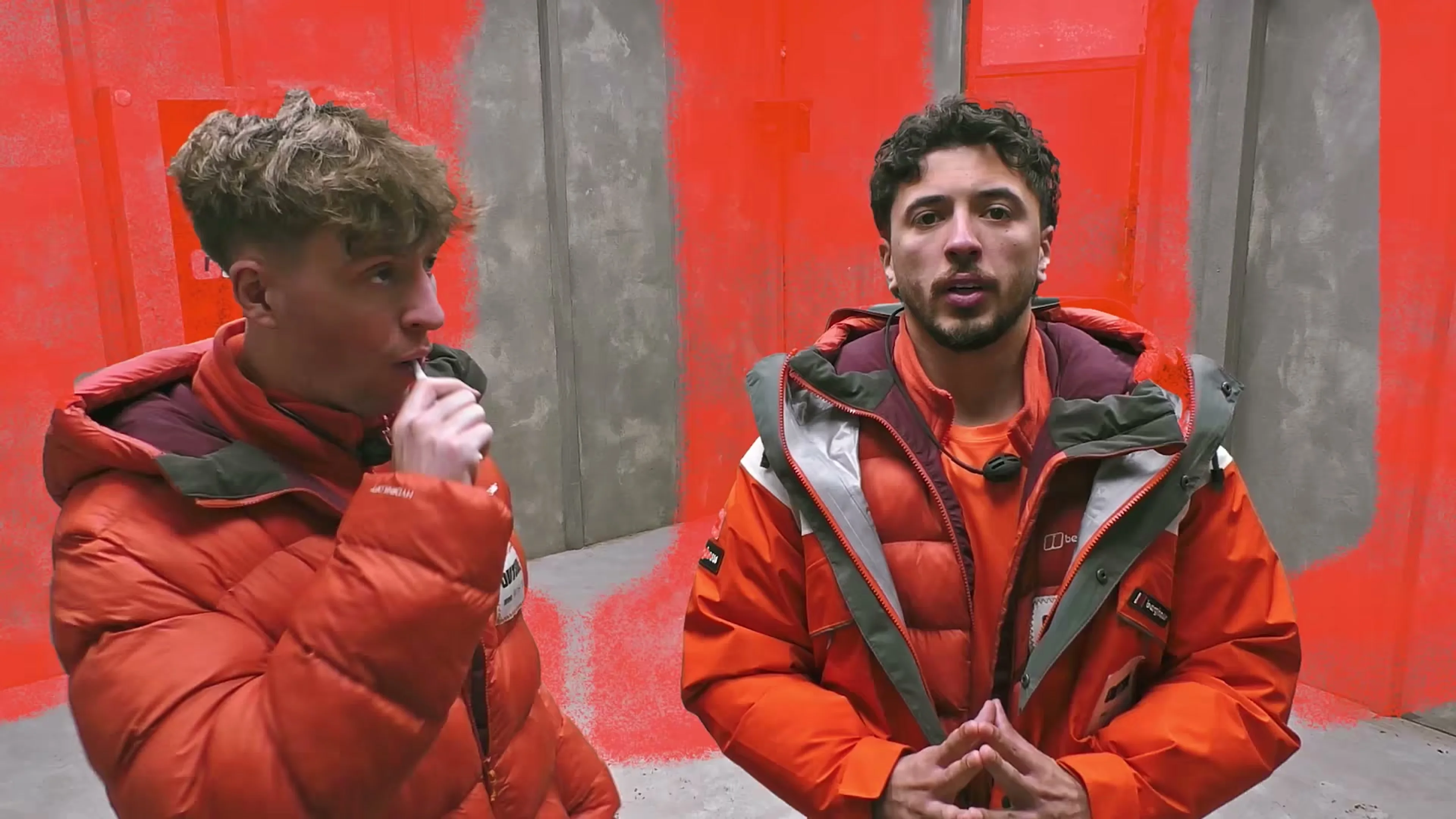 Two contestants in orange jackets talk in a challenge room on Outside.