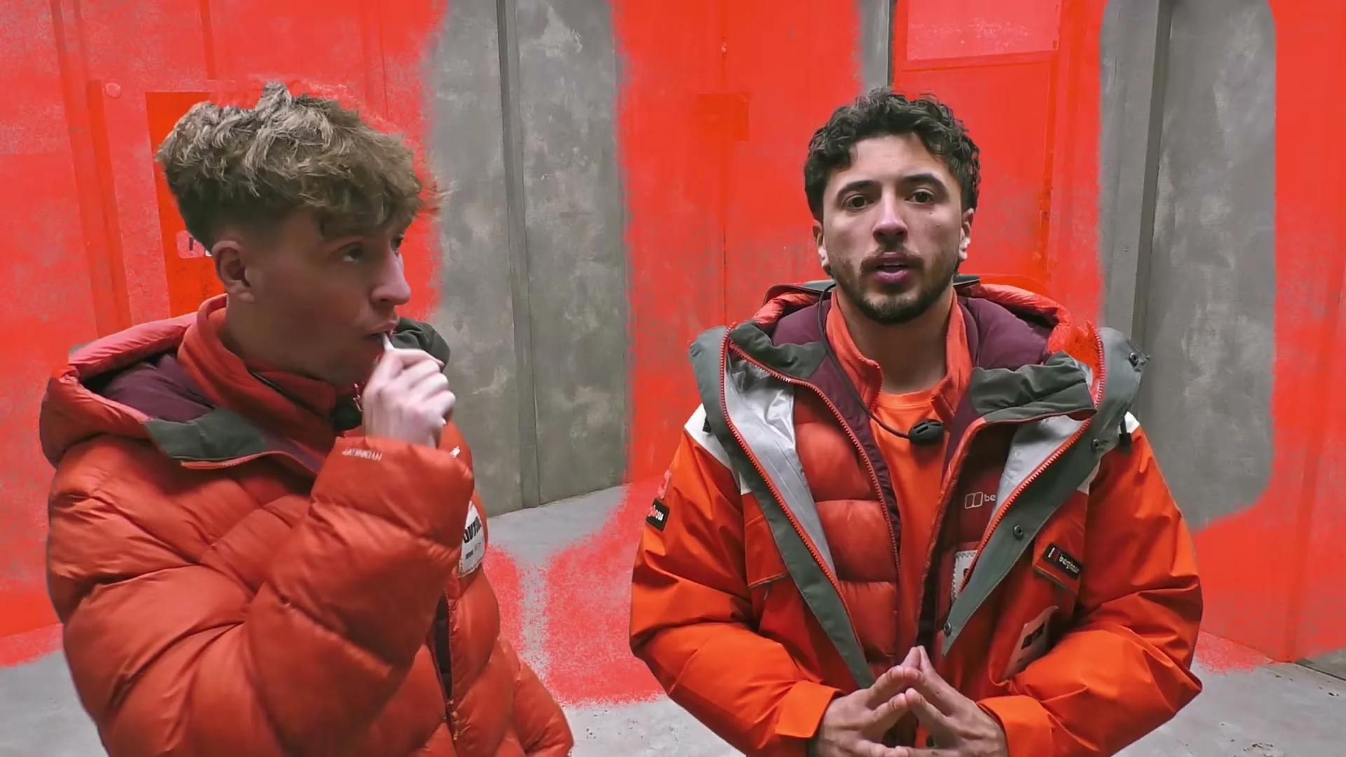 Two contestants in orange jackets talk in a challenge room on Outside.