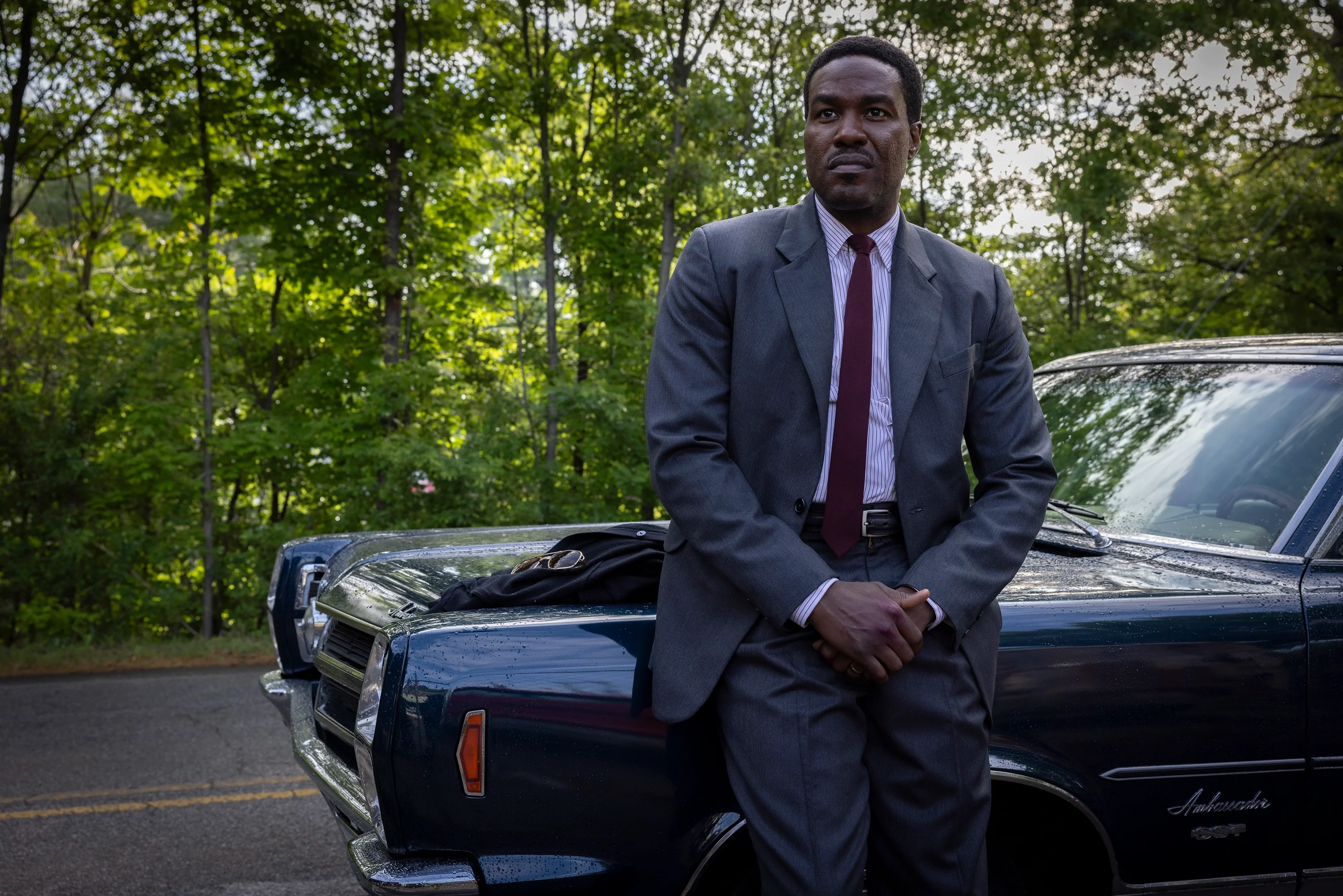 Yahya Abdul-Mateen II as Wayne Strider stands beside a dark blue car in By Any Means.
