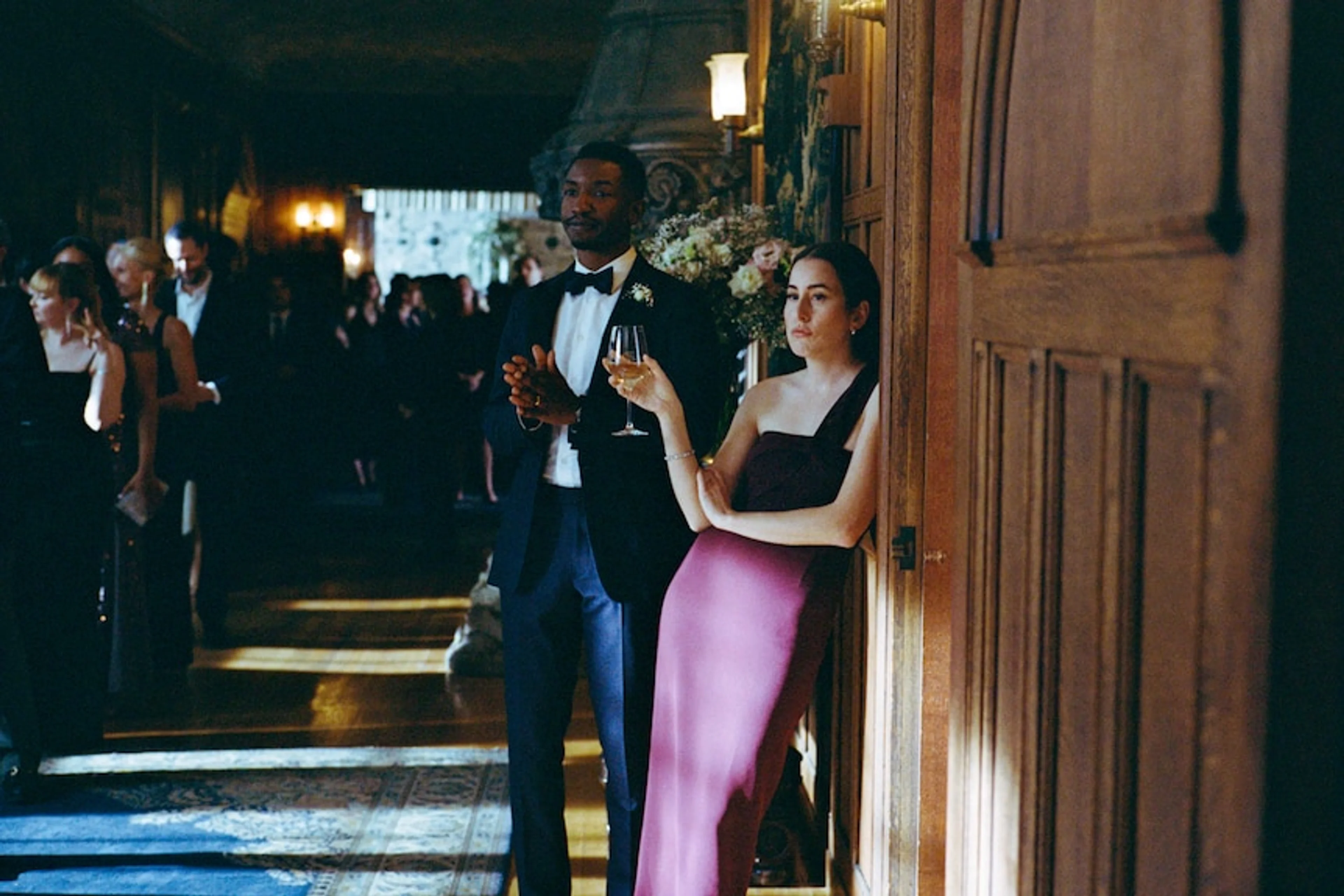 Mike stands in a tuxedo beside Rachel, who holds a wine glass near a wooden doorway.