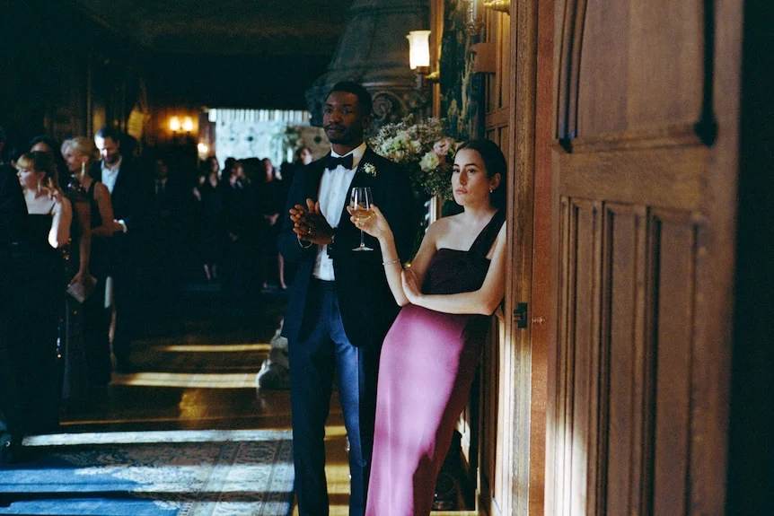 Mike stands in a tuxedo beside Rachel, who holds a wine glass near a wooden doorway.