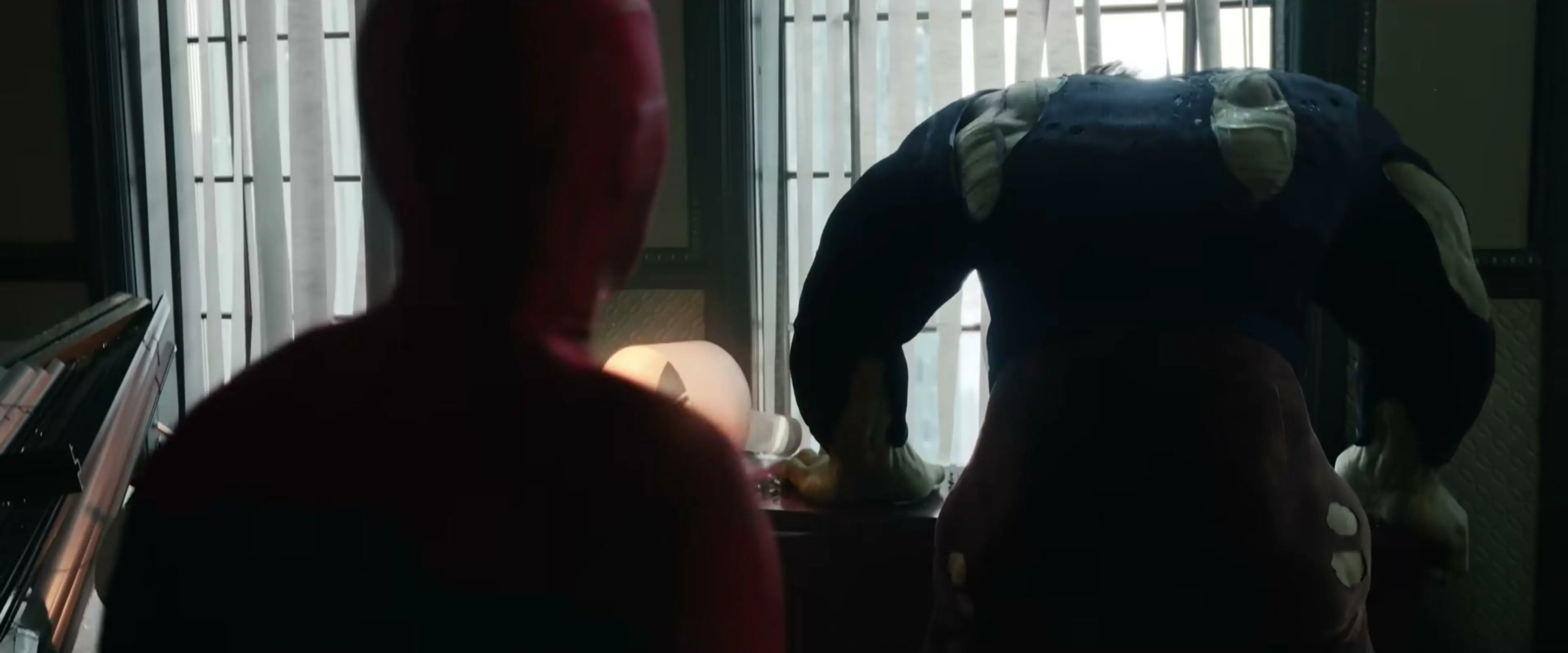 A huge green Hulk bends forward opposite Spider-Man in a room beside vertical window blinds.