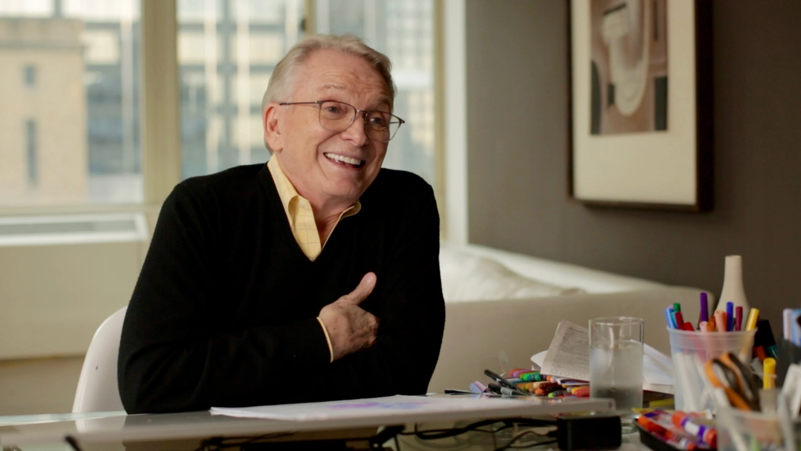 Bob Mackie smiles while seated at a worktable with drawing supplies.