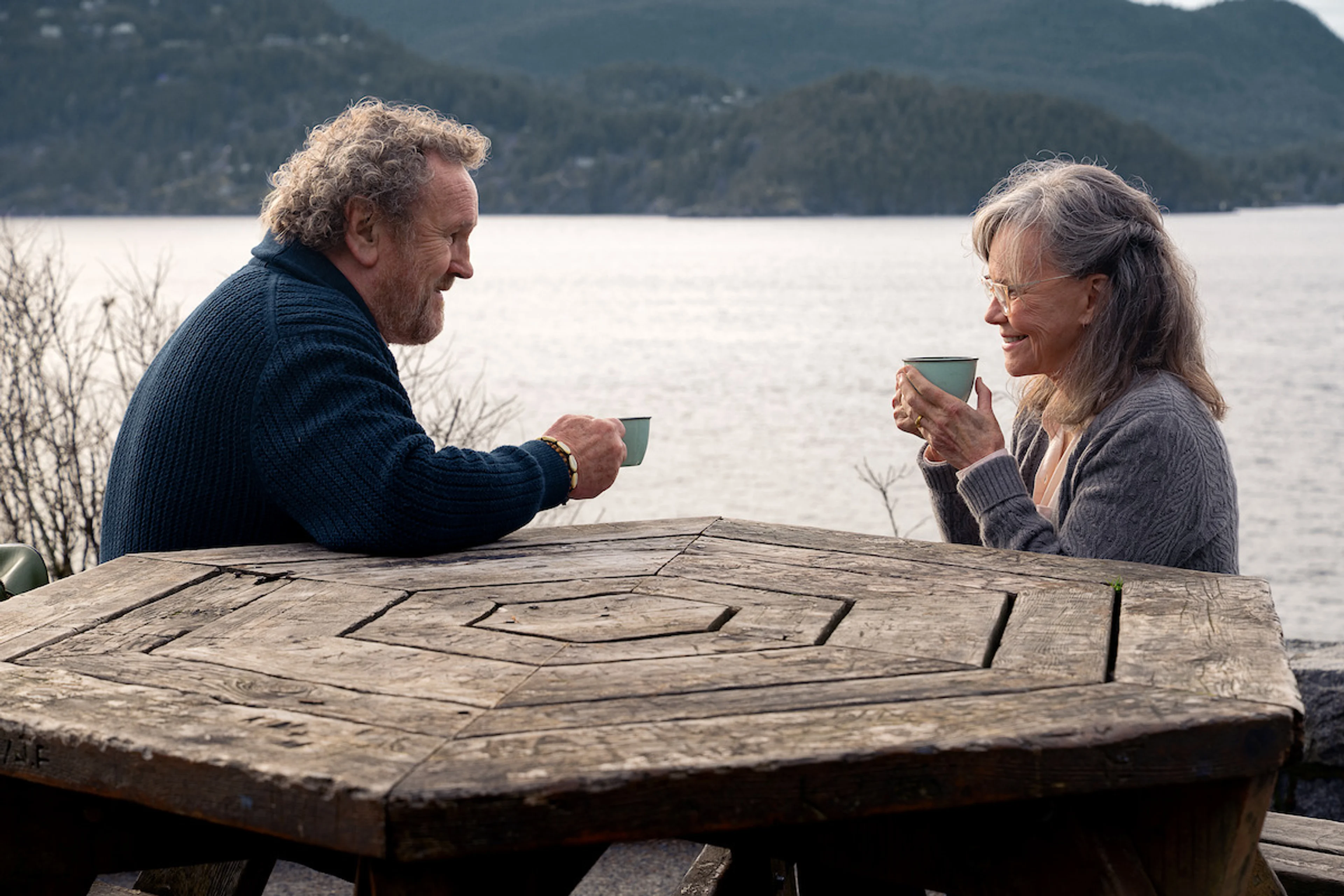 Colm Meaney as Ethan and Sally Field as Tova share hot drinks at a lakeside picnic table.