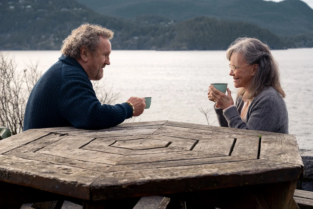 Colm Meaney as Ethan and Sally Field as Tova share hot drinks at a lakeside picnic table.