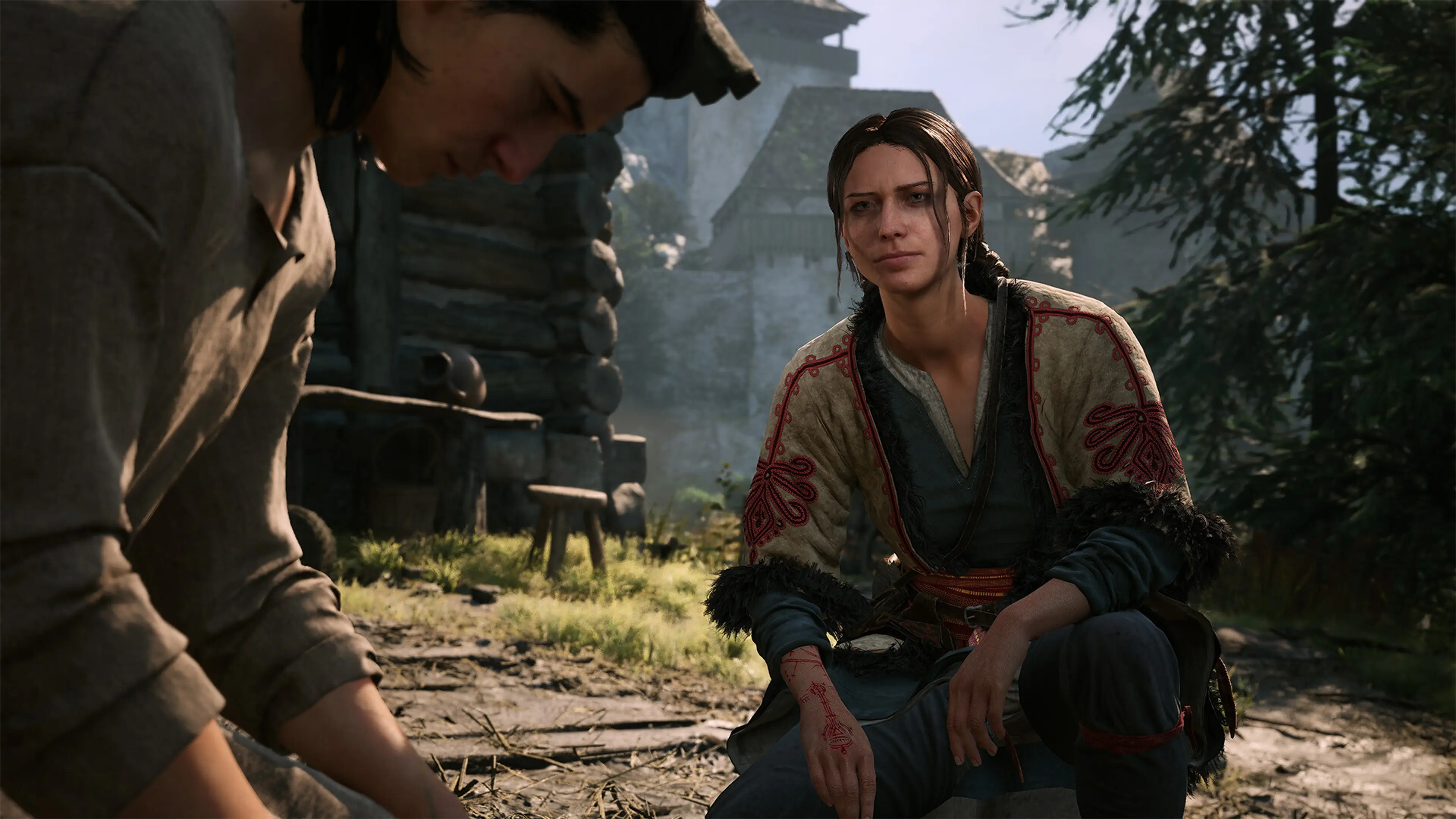 A woman kneels beside another villager outside a rural home in The Blood of Dawnwalker.
