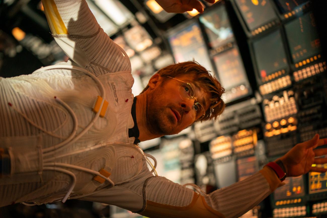 Ryan Gosling floats beside illuminated spacecraft controls with his arms outstretched.