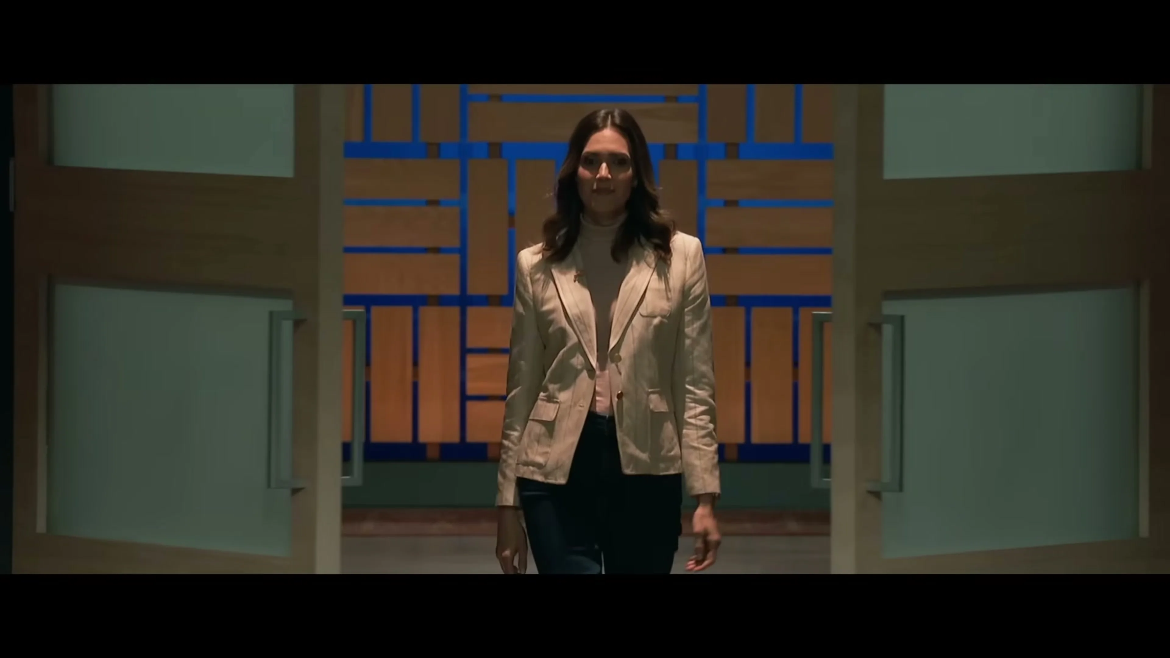 Katie walks through wooden doors wearing a cream jacket.