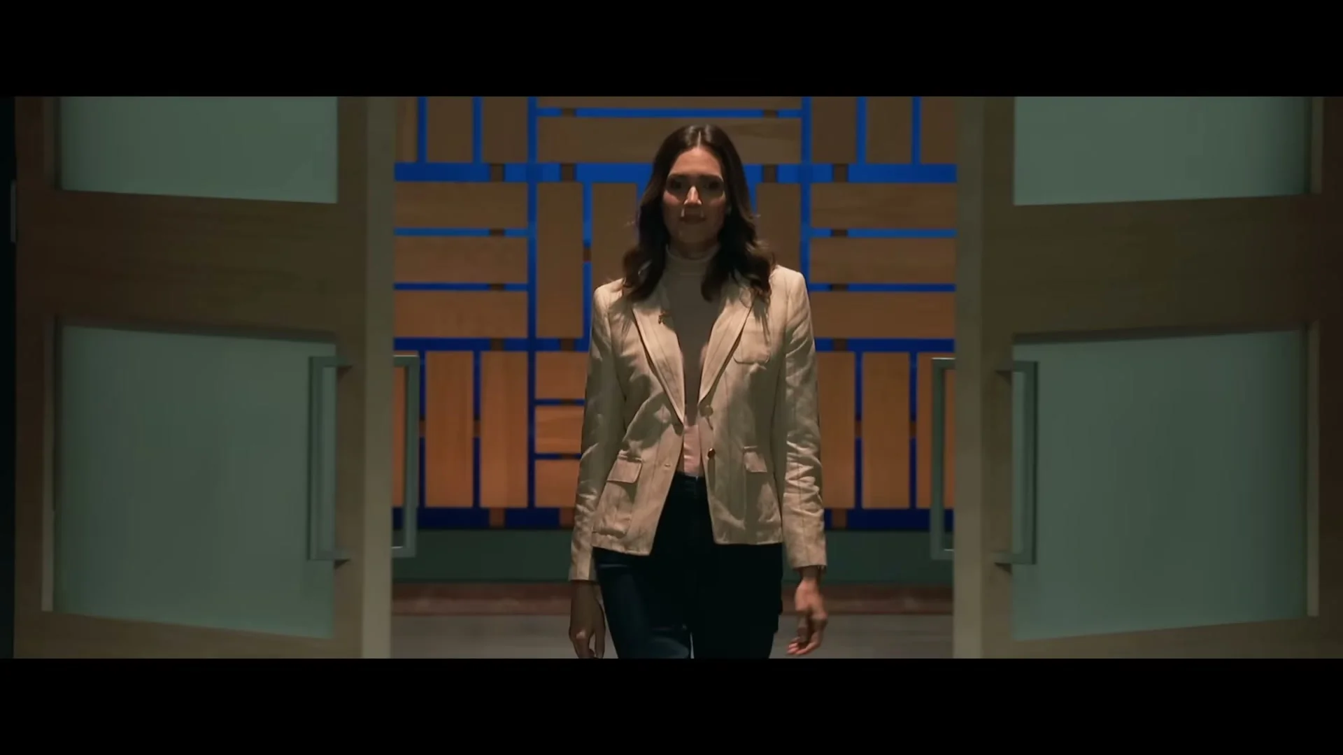Katie walks through wooden doors wearing a cream jacket.