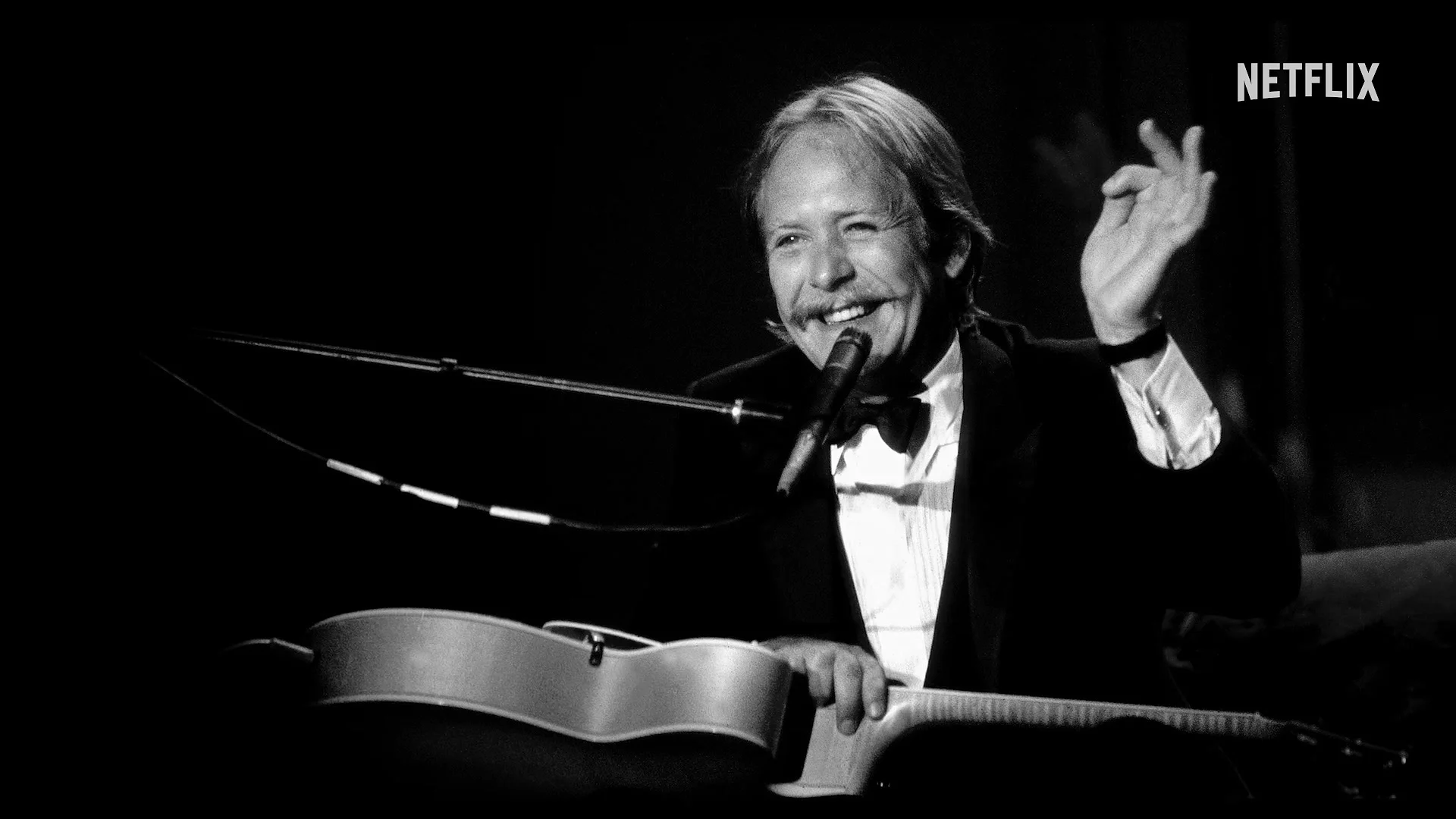 Martin Mull smiles at a microphone with a guitar during an archival performance