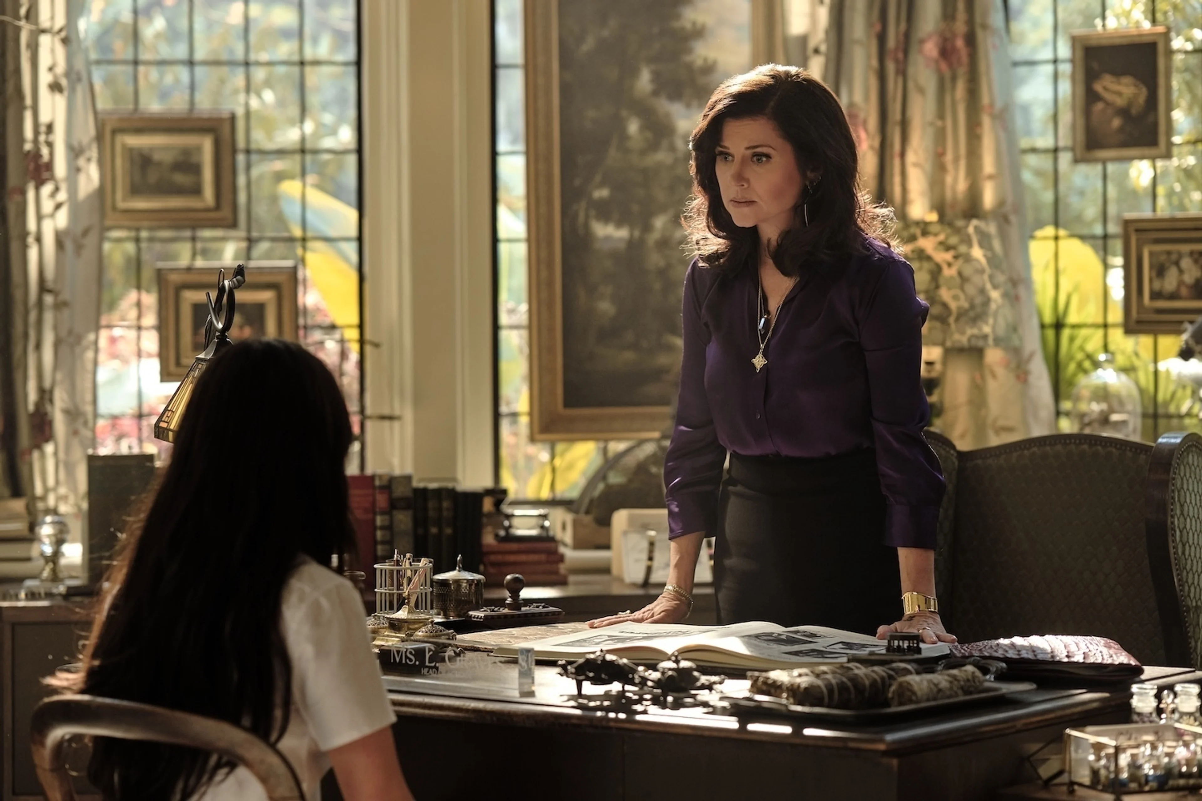 Tiffani Thiessen as Miss Graves stands behind her desk, speaking to a student.