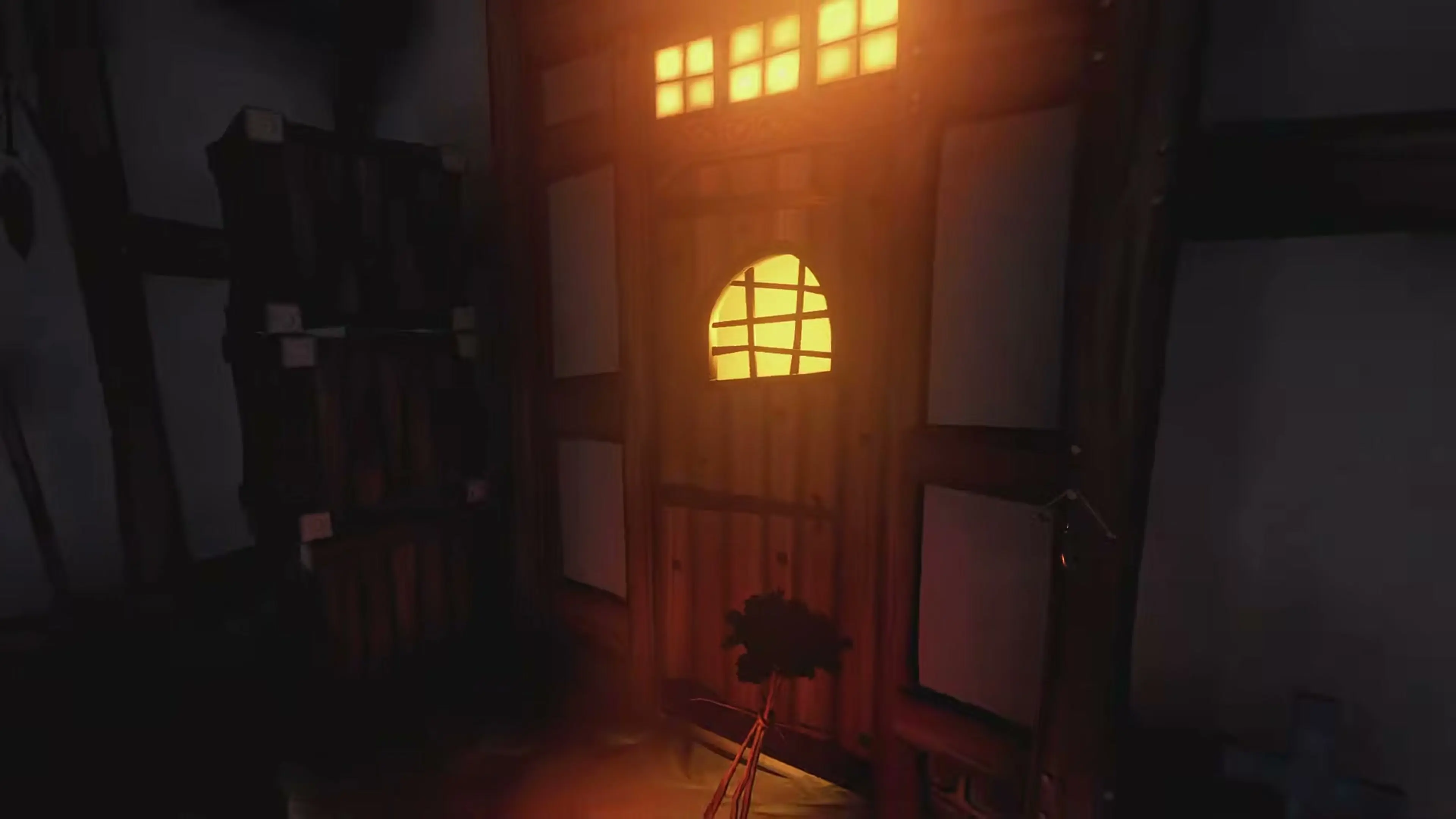 The player hides beside a glowing window and door in the dark village.