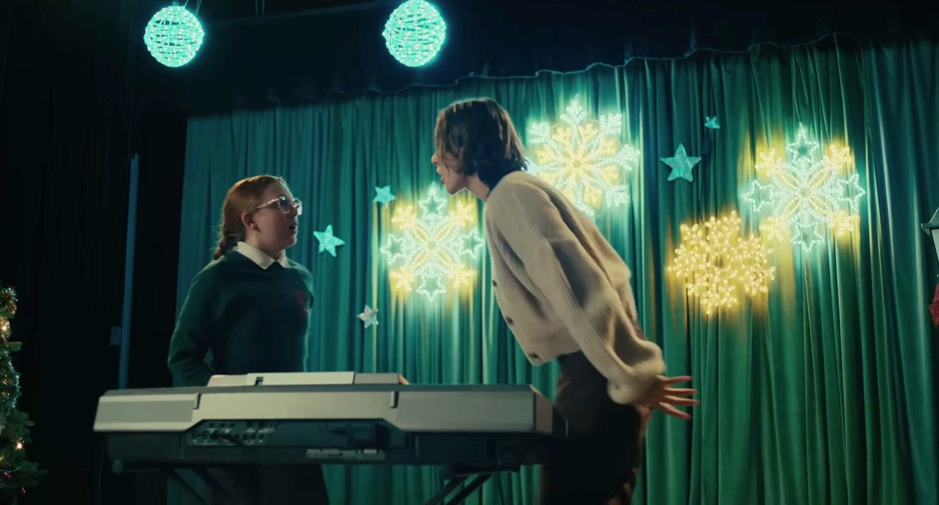 Maria leans toward a pupil beside a keyboard in a school performance scene.