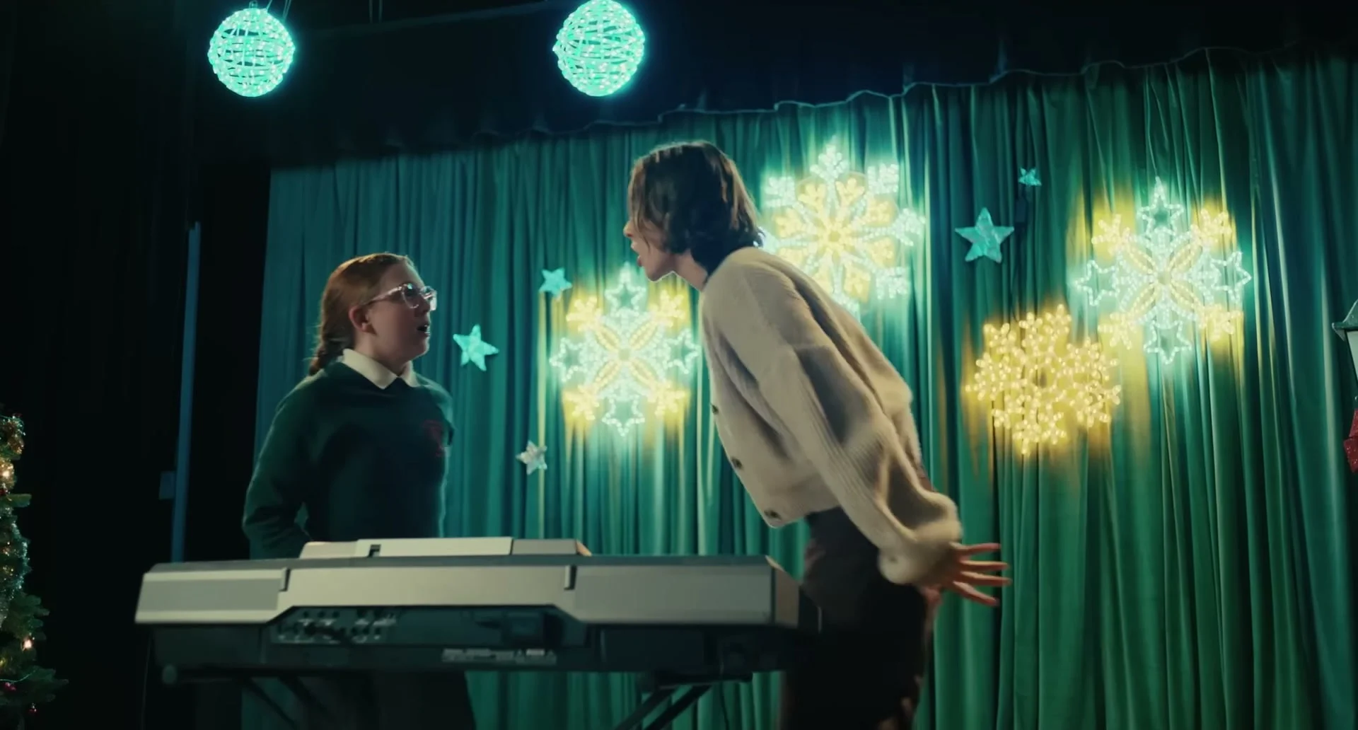 Maria leans toward a pupil beside a keyboard in a school performance scene.