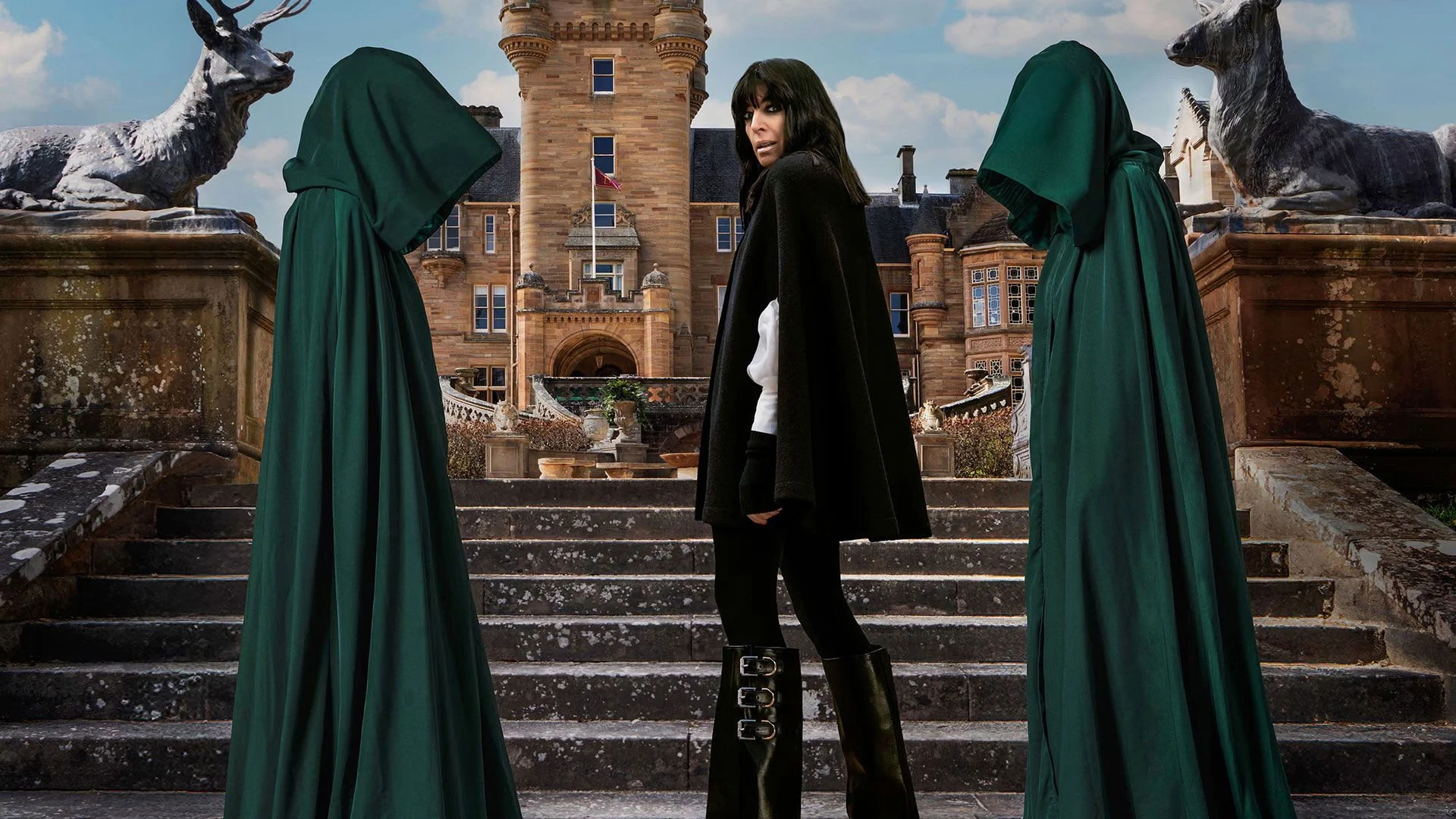 Claudia Winkleman stands on castle steps between two hooded figures for The Celebrity Traitors.