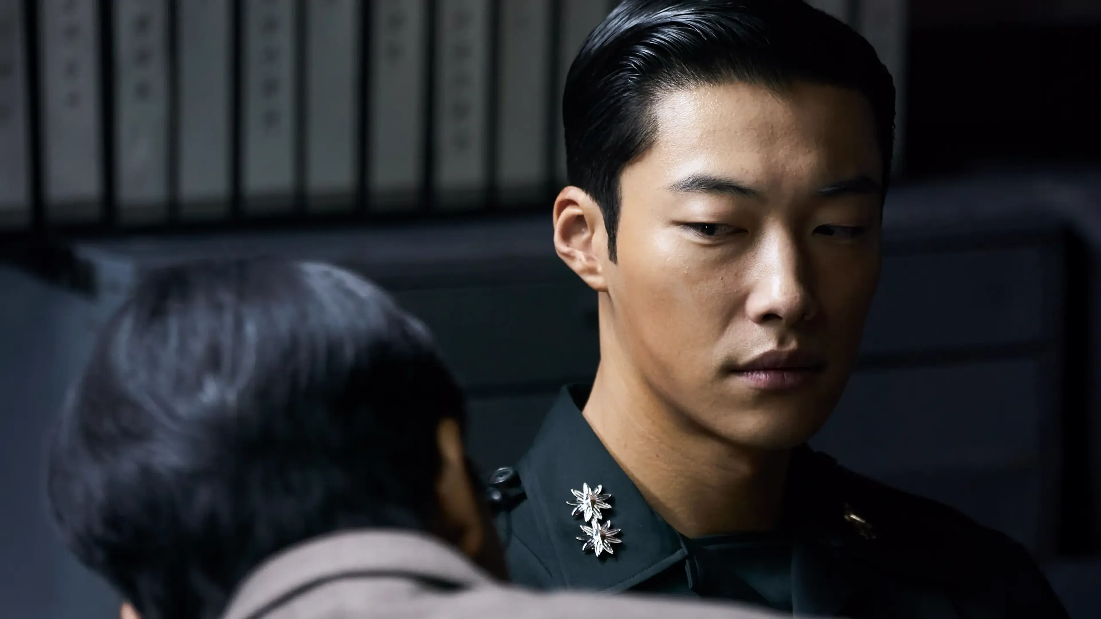 Ki-hyun looks down while wearing a dark military uniform in a dim interior.