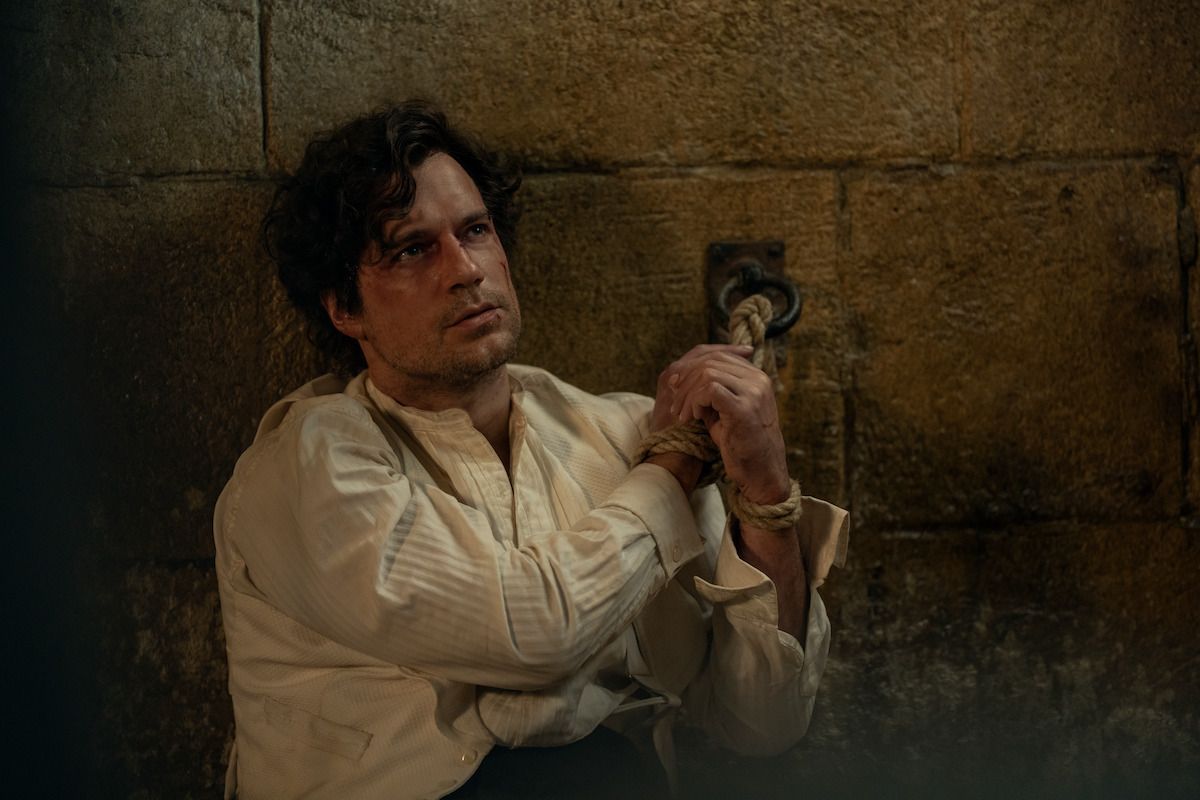 Henry Cavill as Sherlock Holmes sits against a stone wall with his wrists tied.
