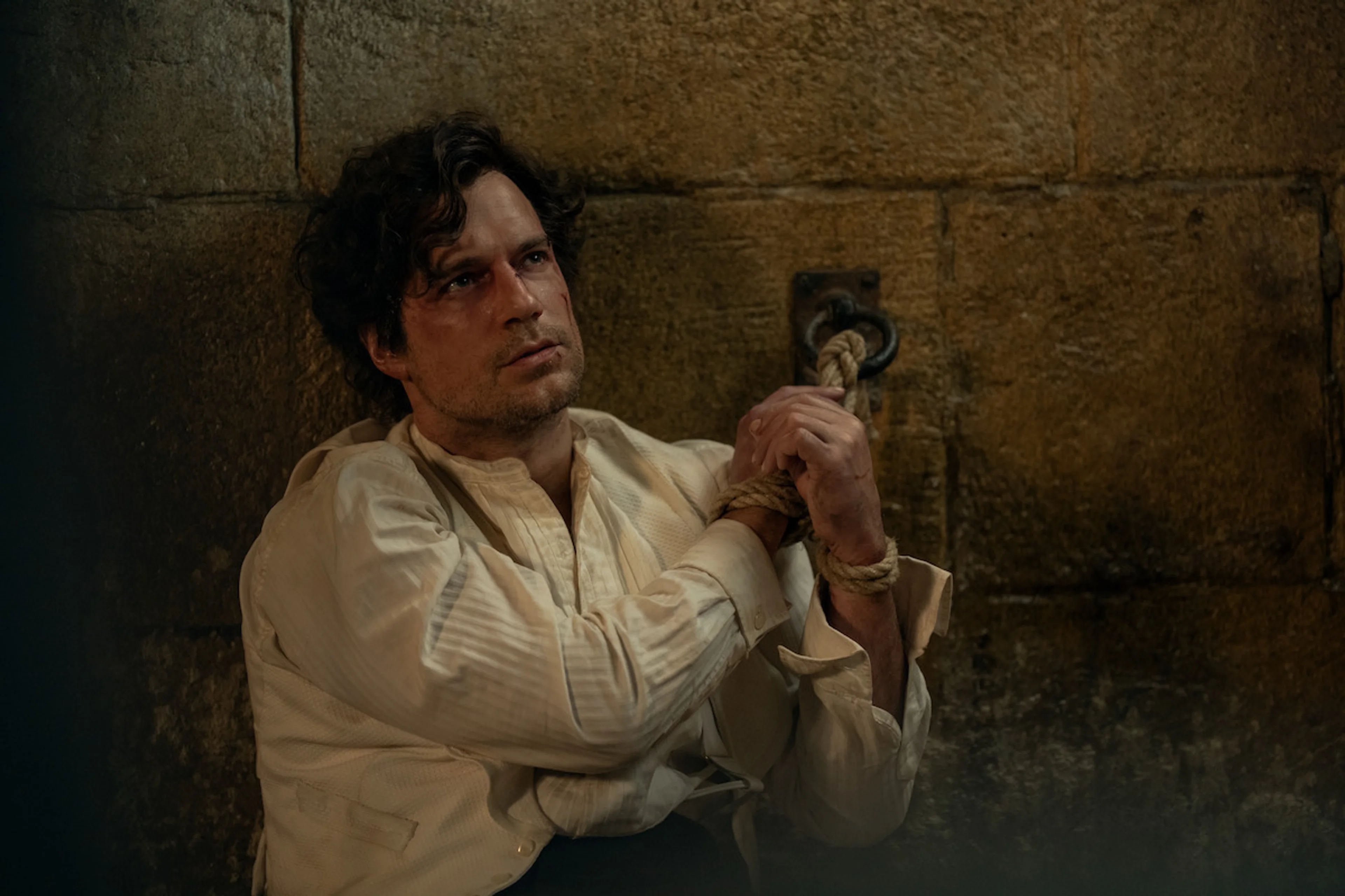 Henry Cavill as Sherlock Holmes sits against a stone wall with his wrists tied.